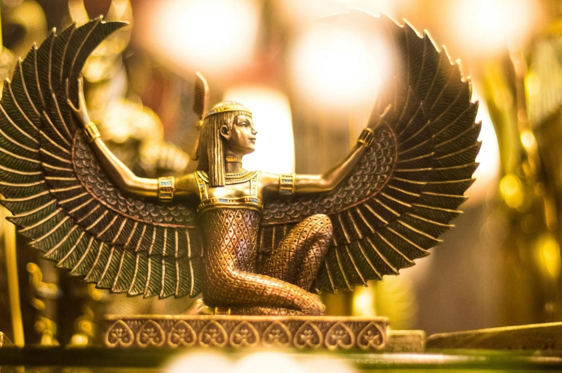 Close-up of a gold-colored statue of a mythological figure with wings, sitting in a crouched position with arms outstretched, detailed with intricate patterns and jewelry, illuminated by warm lighting.