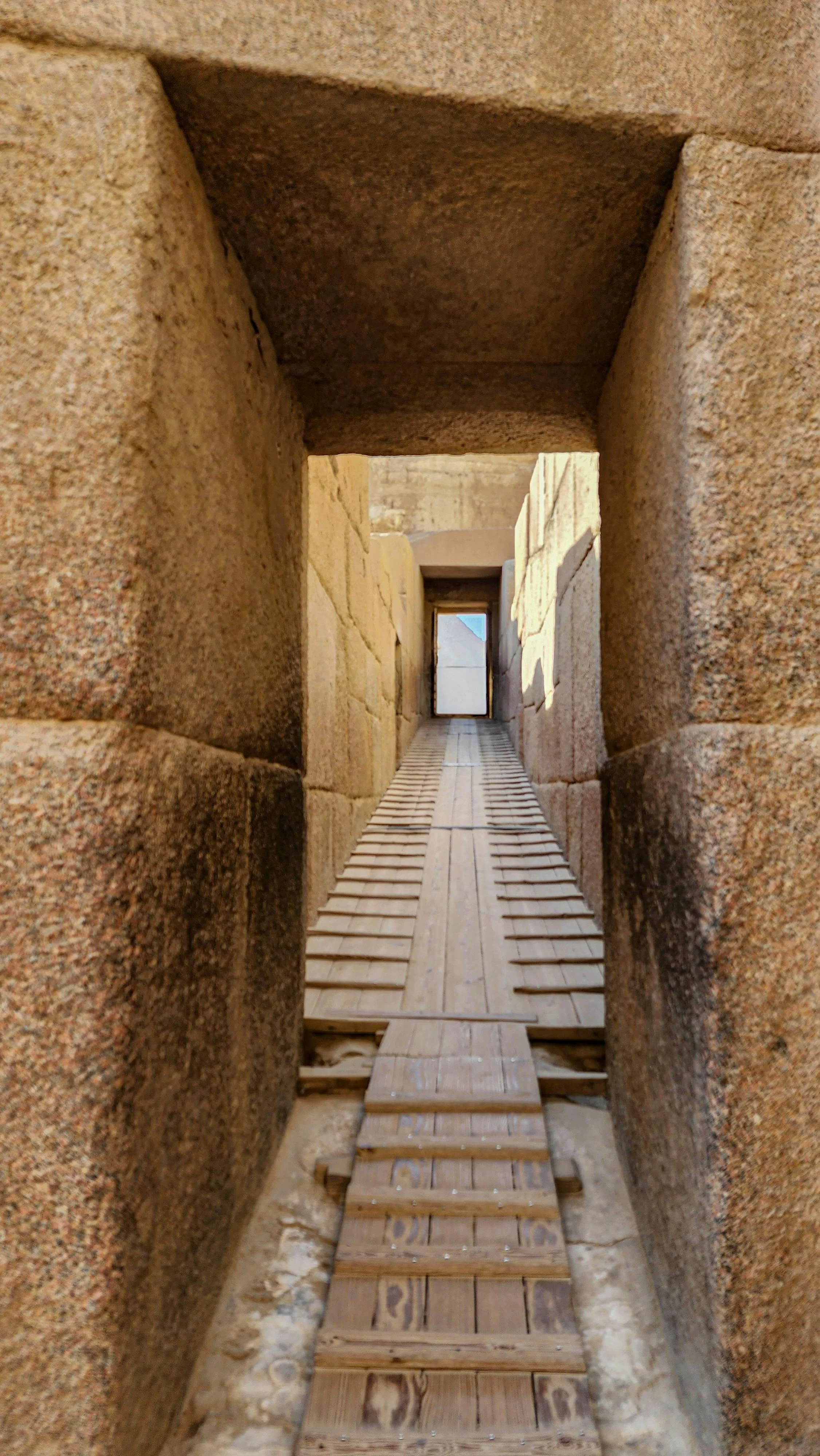 A narrow stone corridor with wooden planks on the floor, leading to an opening with sky visible beyond.