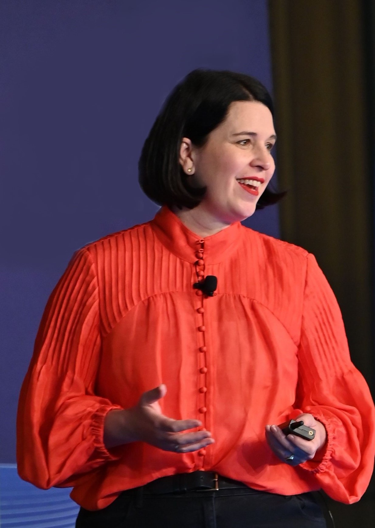 Karlee, a woman with dark hair, is wearing a bright red blouse, speaking at a conference standing on stage against a dark purple background.
