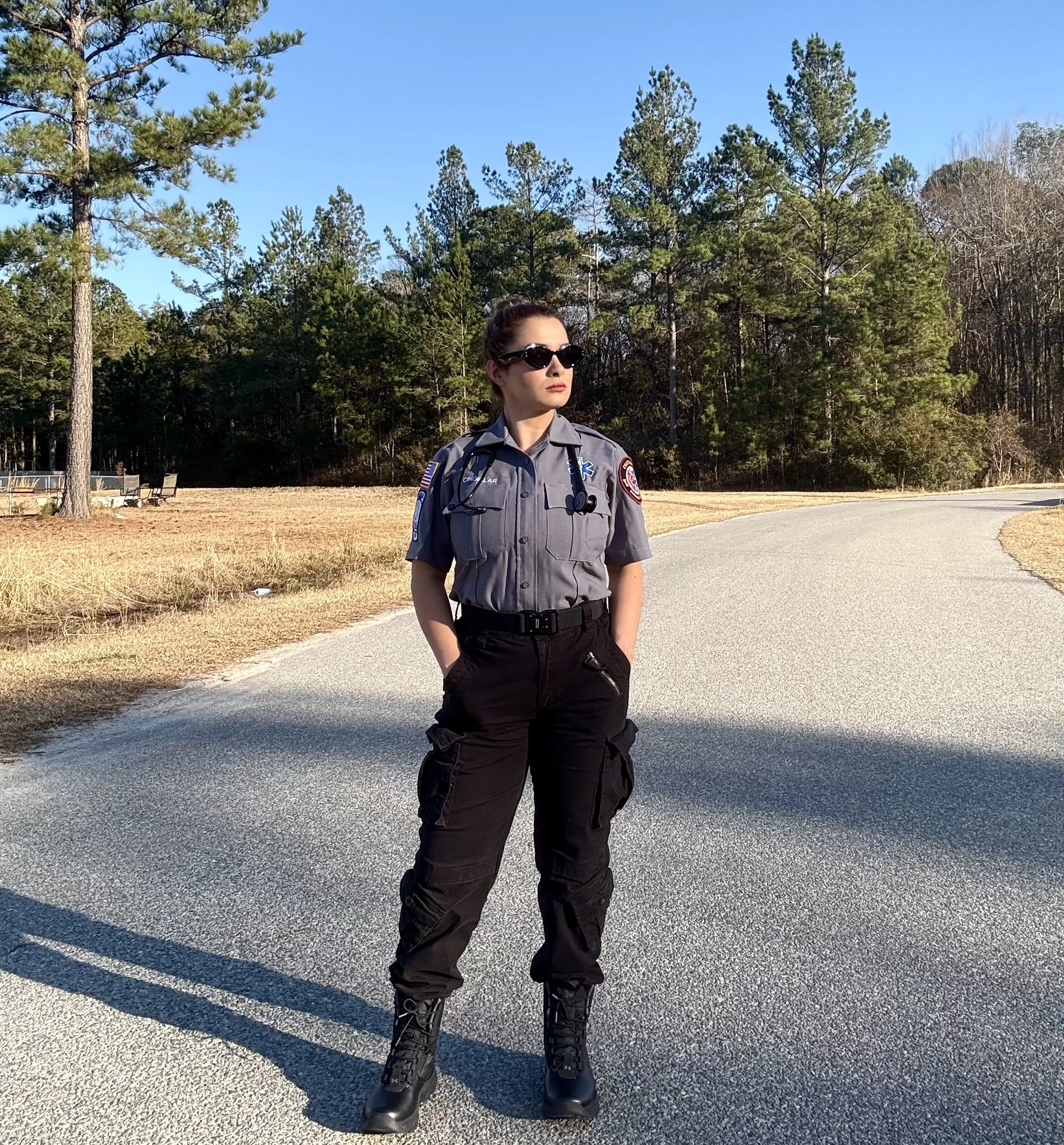 A female paramedic in uniform standing on a paved path in a park with tall trees and blue sky in the background.