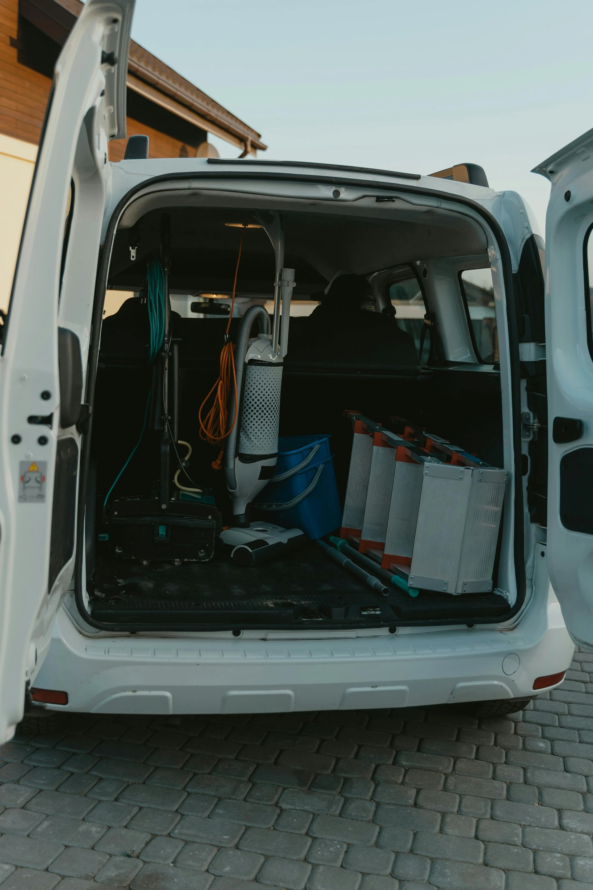 Open van trunk filled with cleaning equipment like ladders, mop, vacuum, and cleaning supplies, parked on a paved driveway.