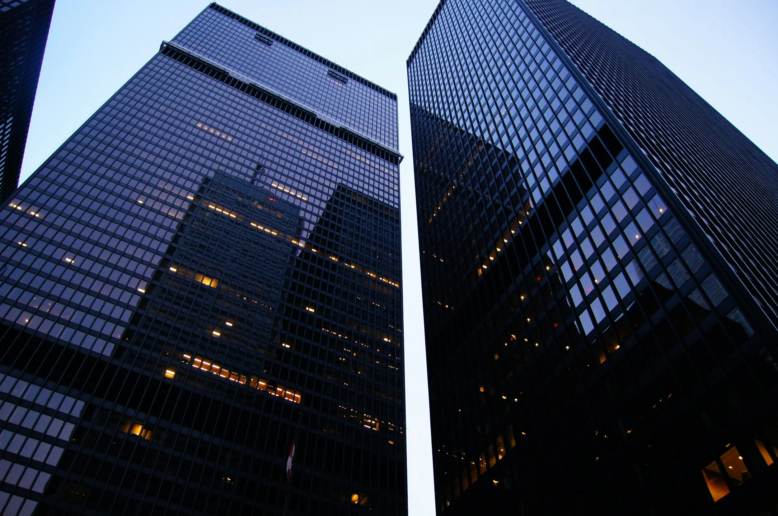 Two tall glass skyscrapers reflect the sky and surrounding buildings, seen from ground level looking upwards.
