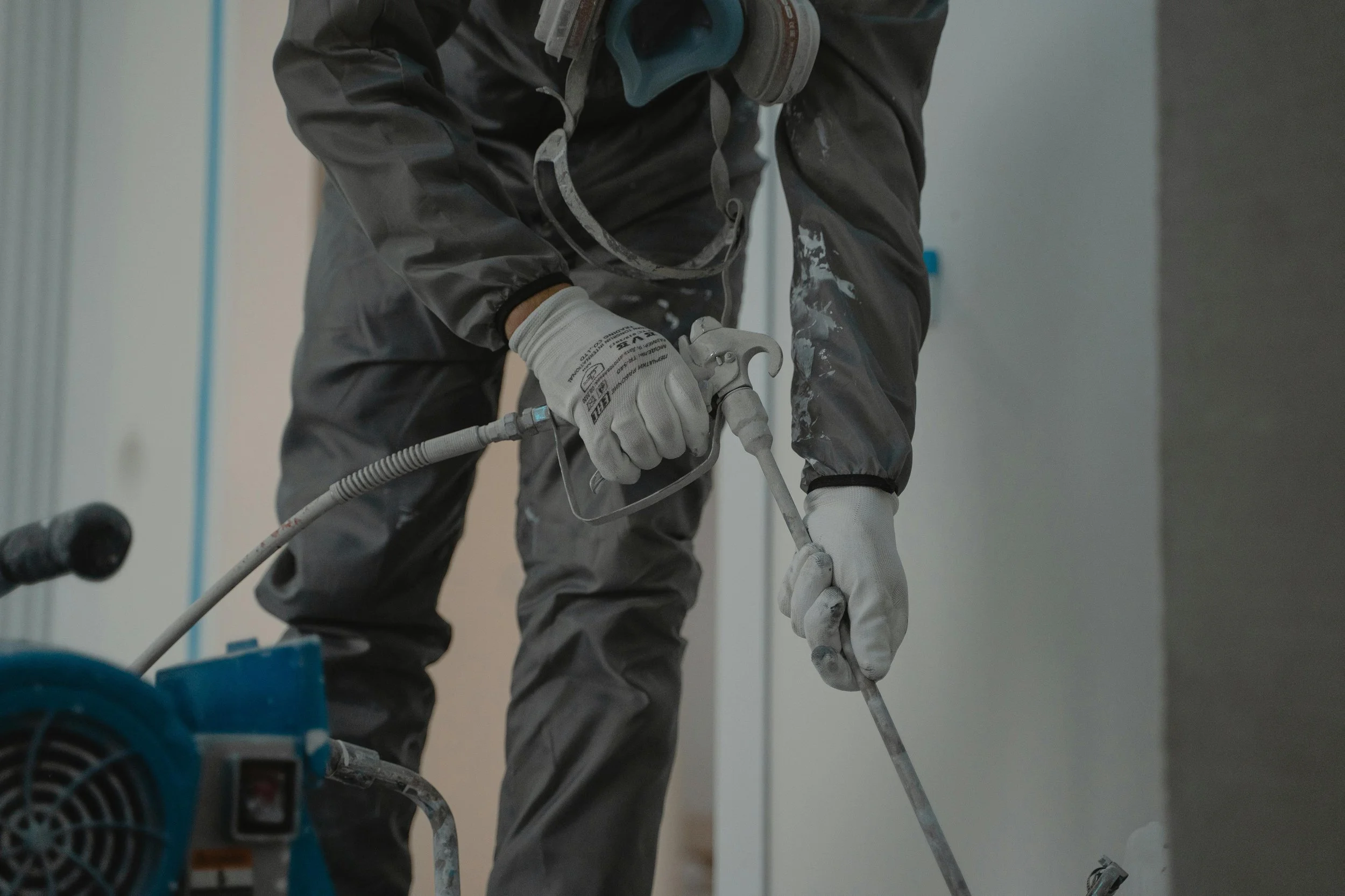 A worker in protective clothing and gloves uses a power tool to sand or grind a wall in an indoor construction or renovation site.