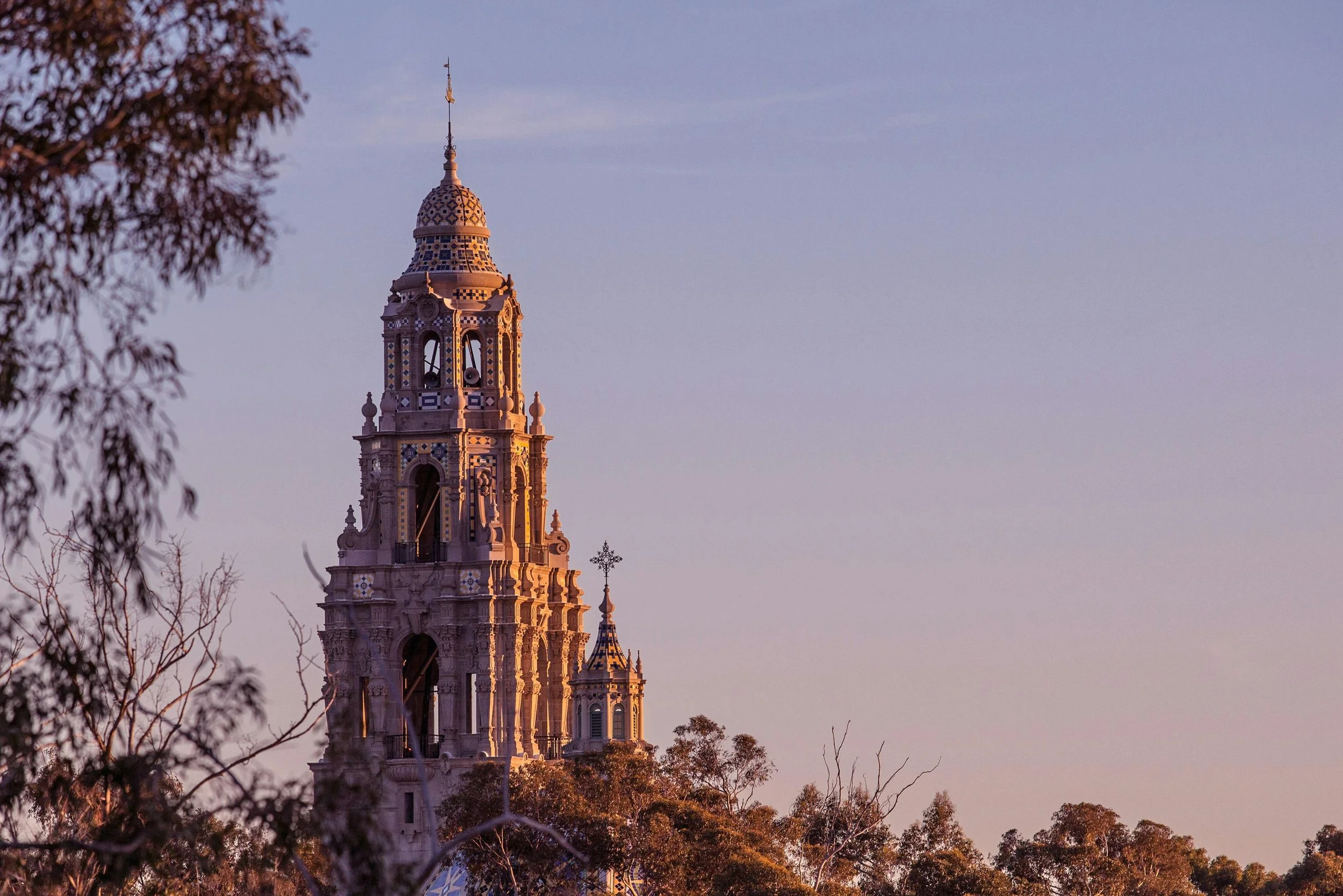 A tall, ornate tower with intricate architectural details stands against a soft purple sky, surrounded by trees.
