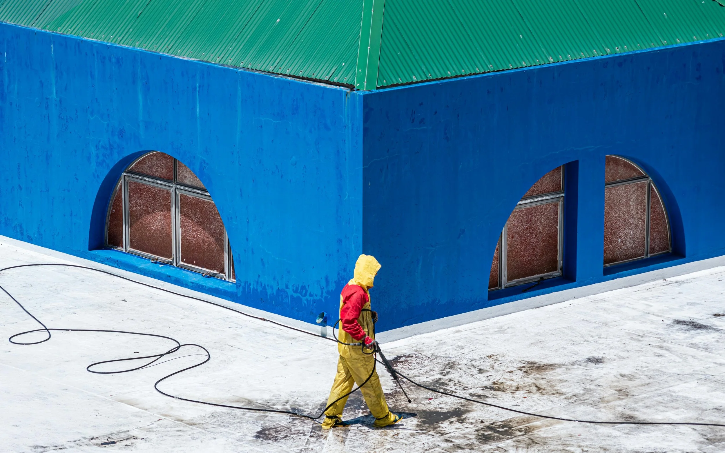 Worker in yellow protective suit pressure washing a white rooftop with a blue building in the background.