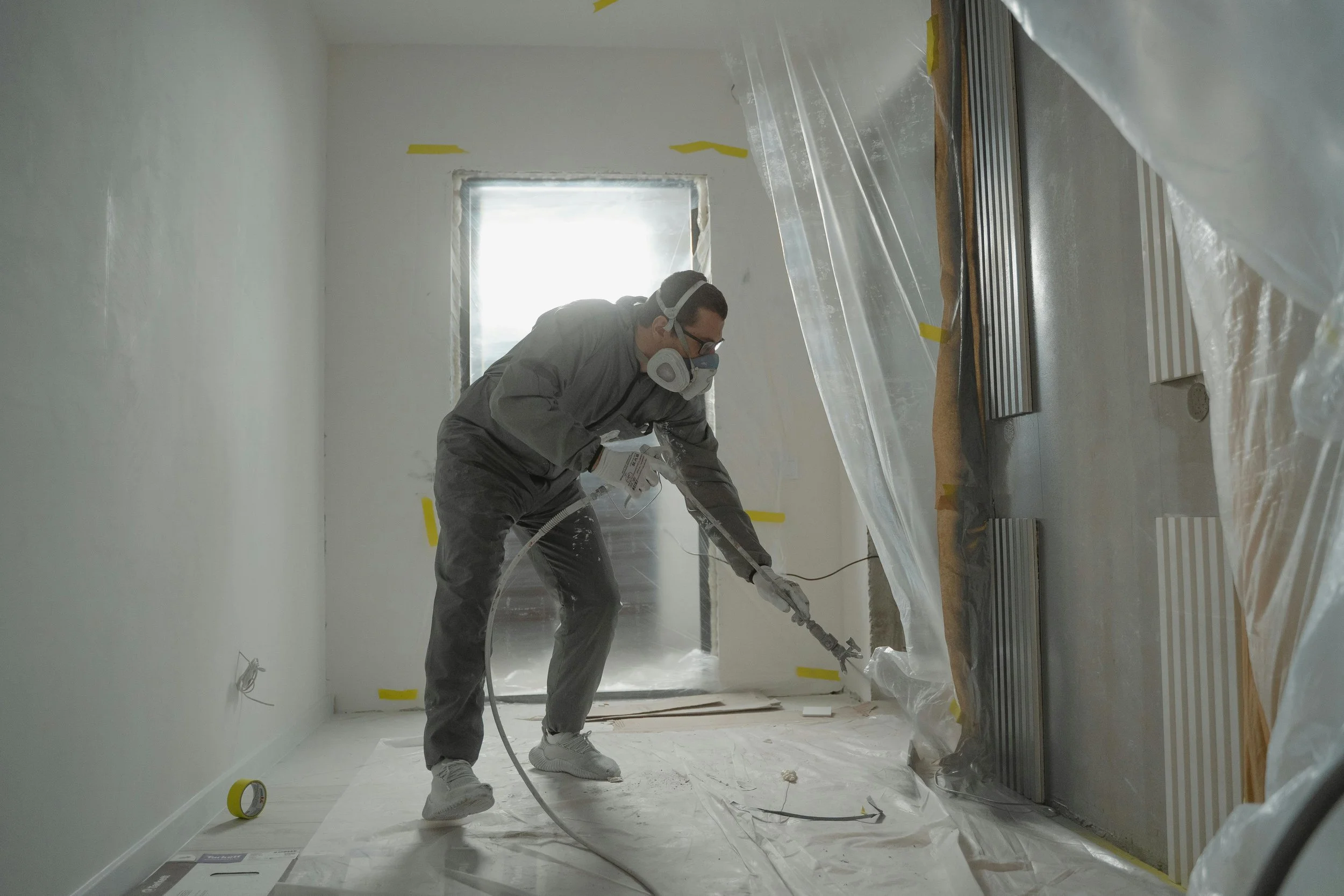 A person wearing protective gear, mask, and gloves using a spray gun to paint a wall in a room under construction or renovation.