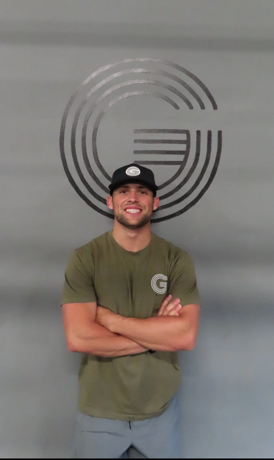 A young man smiling with arms crossed in front of a gray wall with a large circular logo. He is wearing a black cap and an olive green t-shirt, both featuring a logo with a stylized letter 'G' and concentric oval lines.