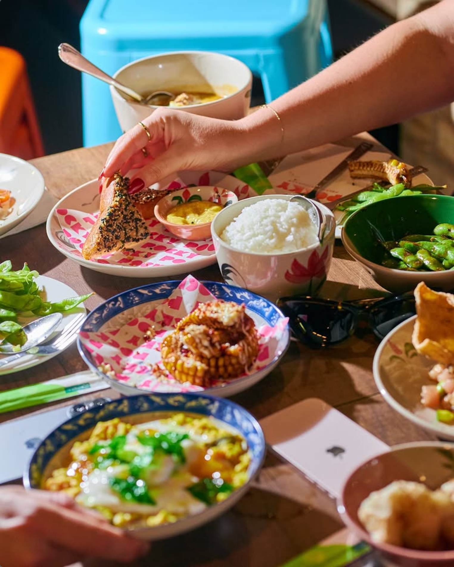 A table filled with various dishes including rice, soup, vegetables, and grilled corn along with a person's hand reaching for a piece of bread.