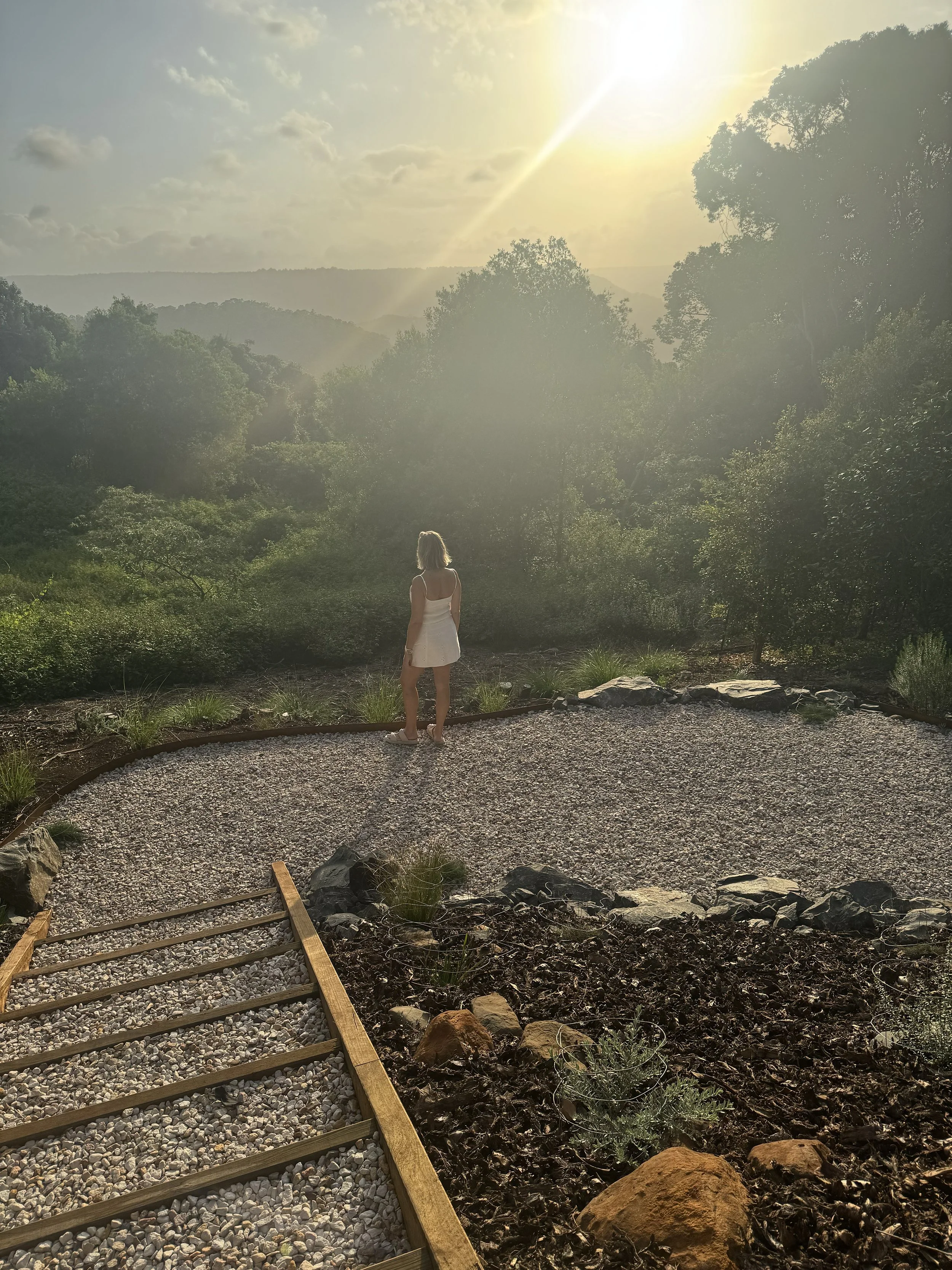 A woman in a white dress standing on a gravel path, looking over a lush, green landscape of trees and hills at sunset.