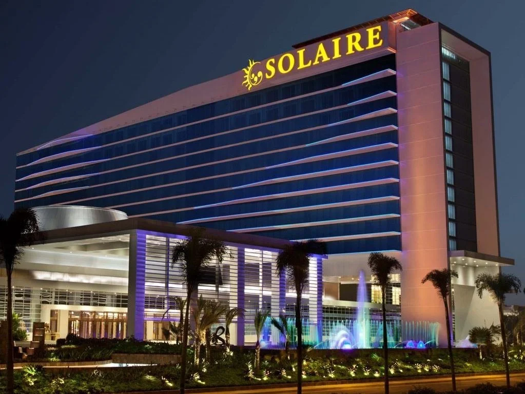 Night view of the Solaire Resort & Casino with illuminated building, palm trees, and colorful fountains.