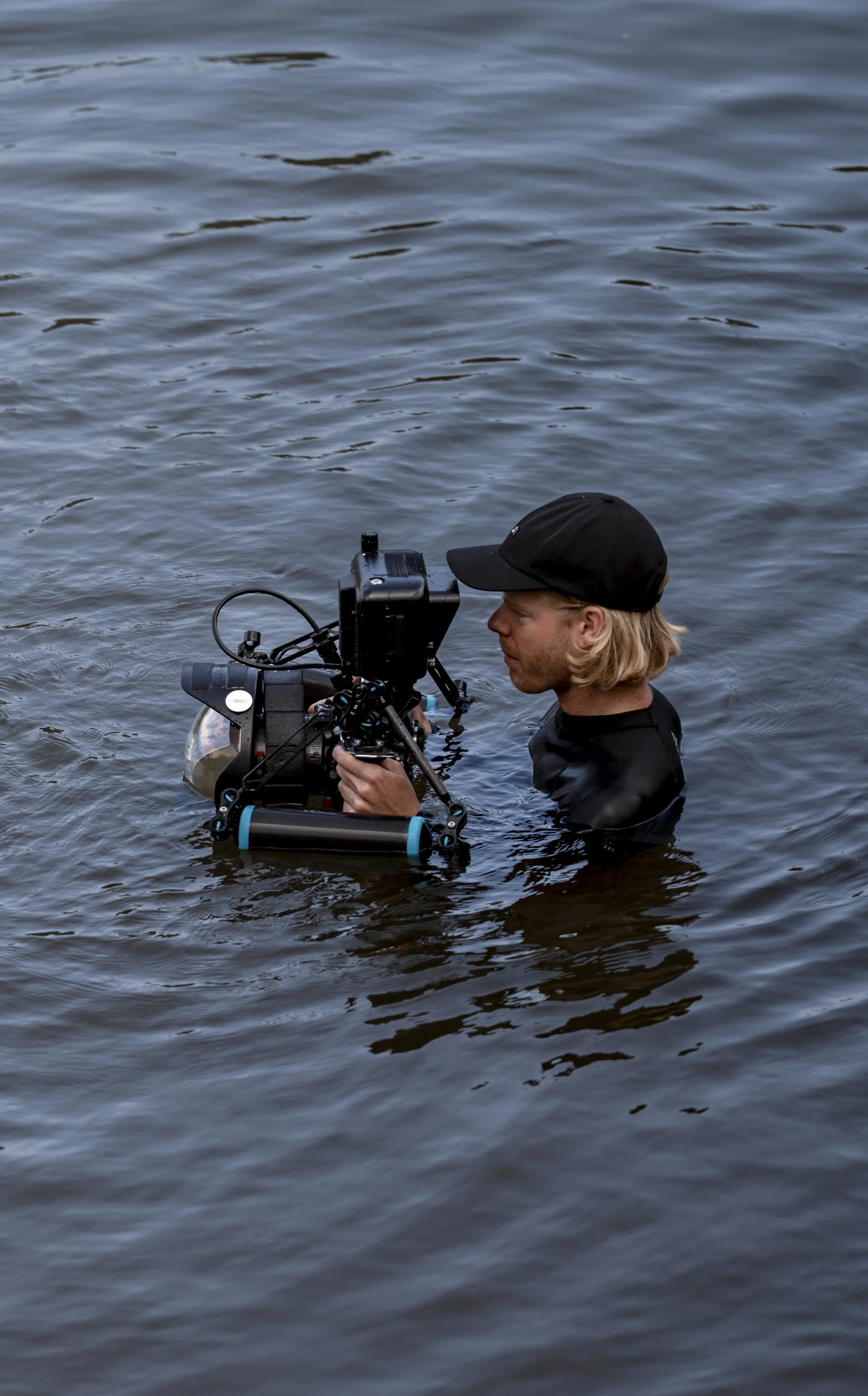 A person with blonde hair, wearing a black cap and black shirt, is standing in water and operating underwater camera equipment.