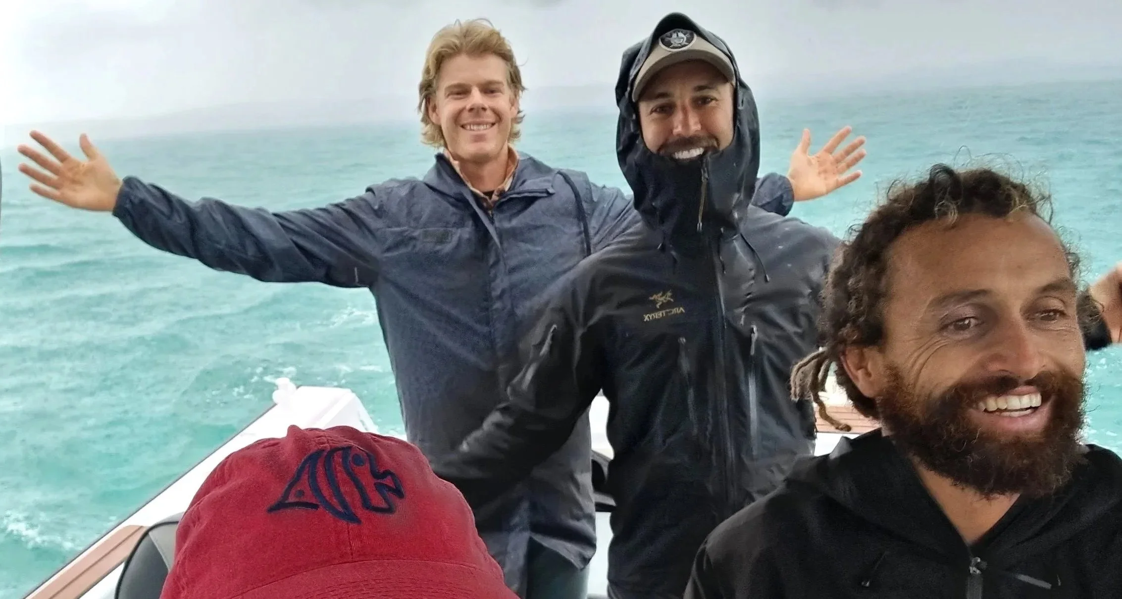 Three men on a boat smiling, with water and cloudy sky in the background, one with arms outstretched.