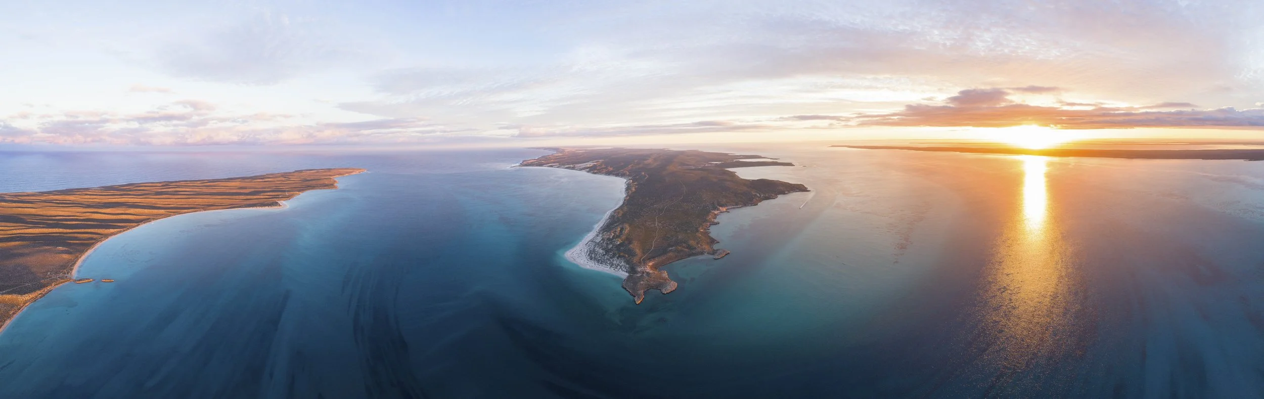Aerial view of a coastal landscape at sunset showing a peninsula, ocean, and sky with clouds.