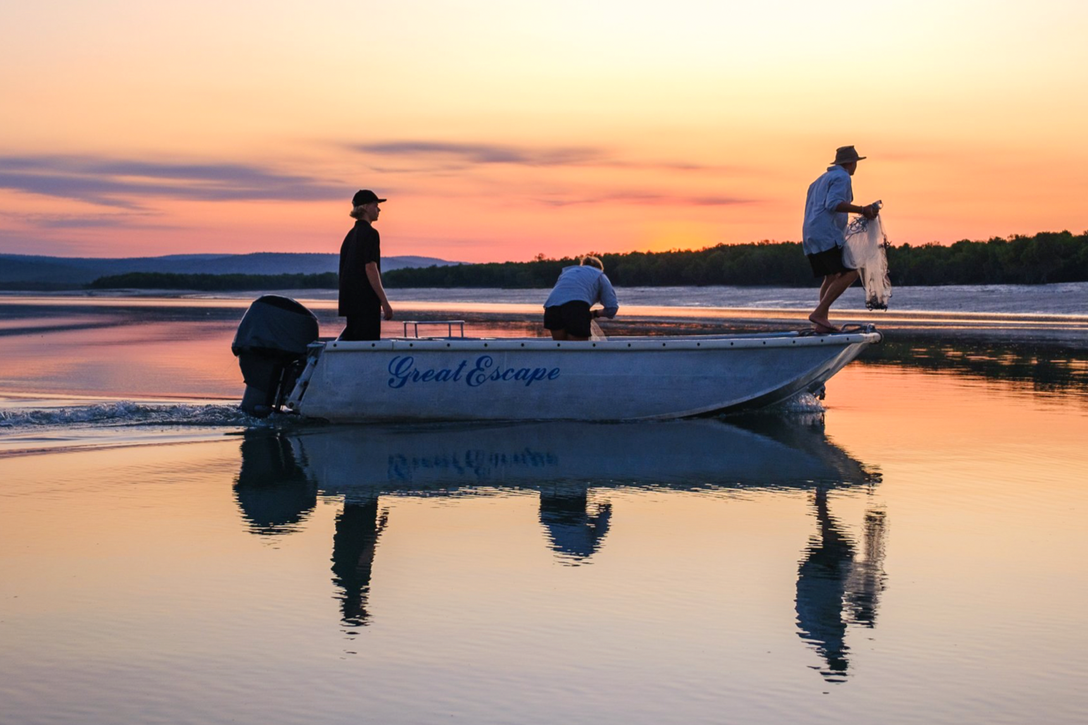 Three people on a boat named 'Great Escape' at sunset, with one standing at the front and two others working near the middle and back, on calm water reflecting the colorful sky.