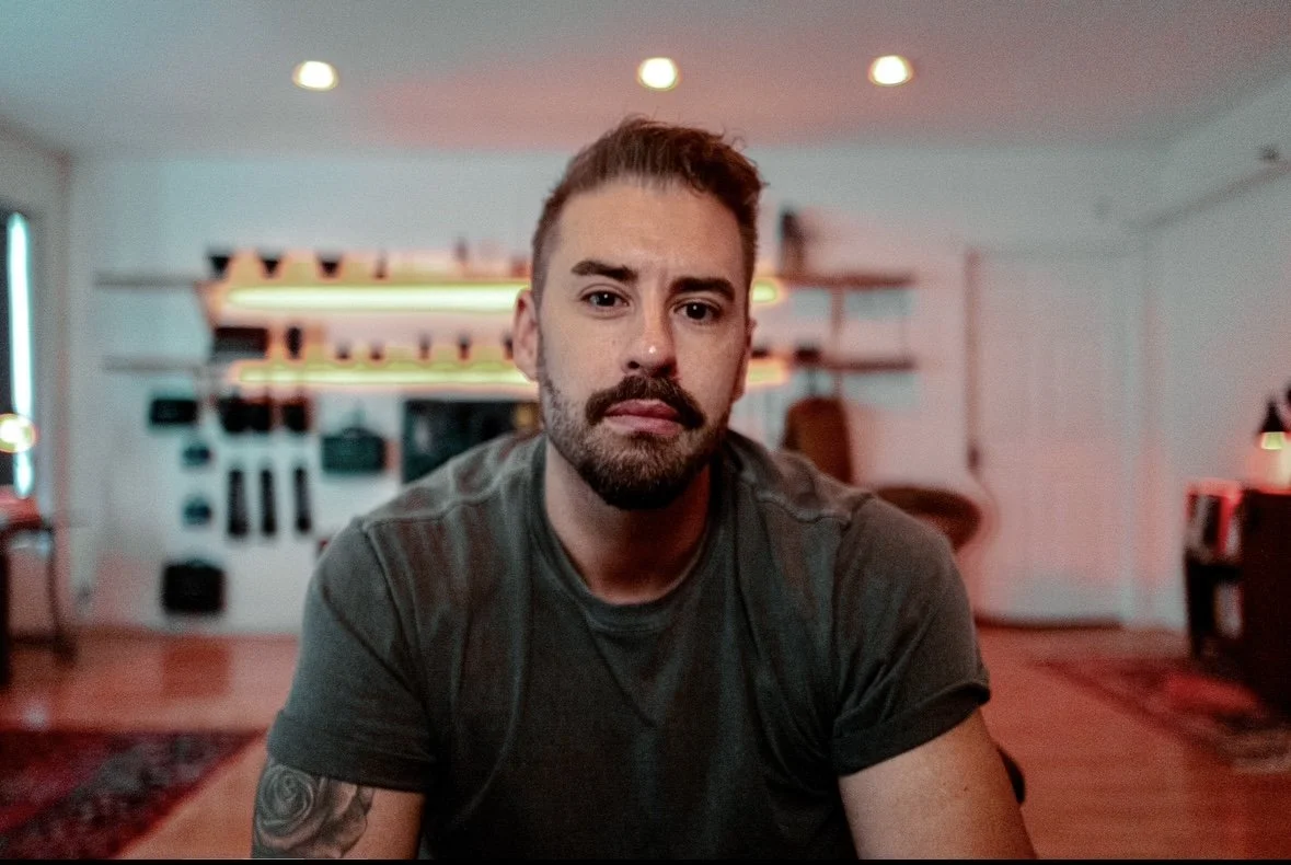 A man with a beard and mustache sitting in a living room, looking into the camera with a neutral expression. There are shelves with objects and a neon sign in the background.