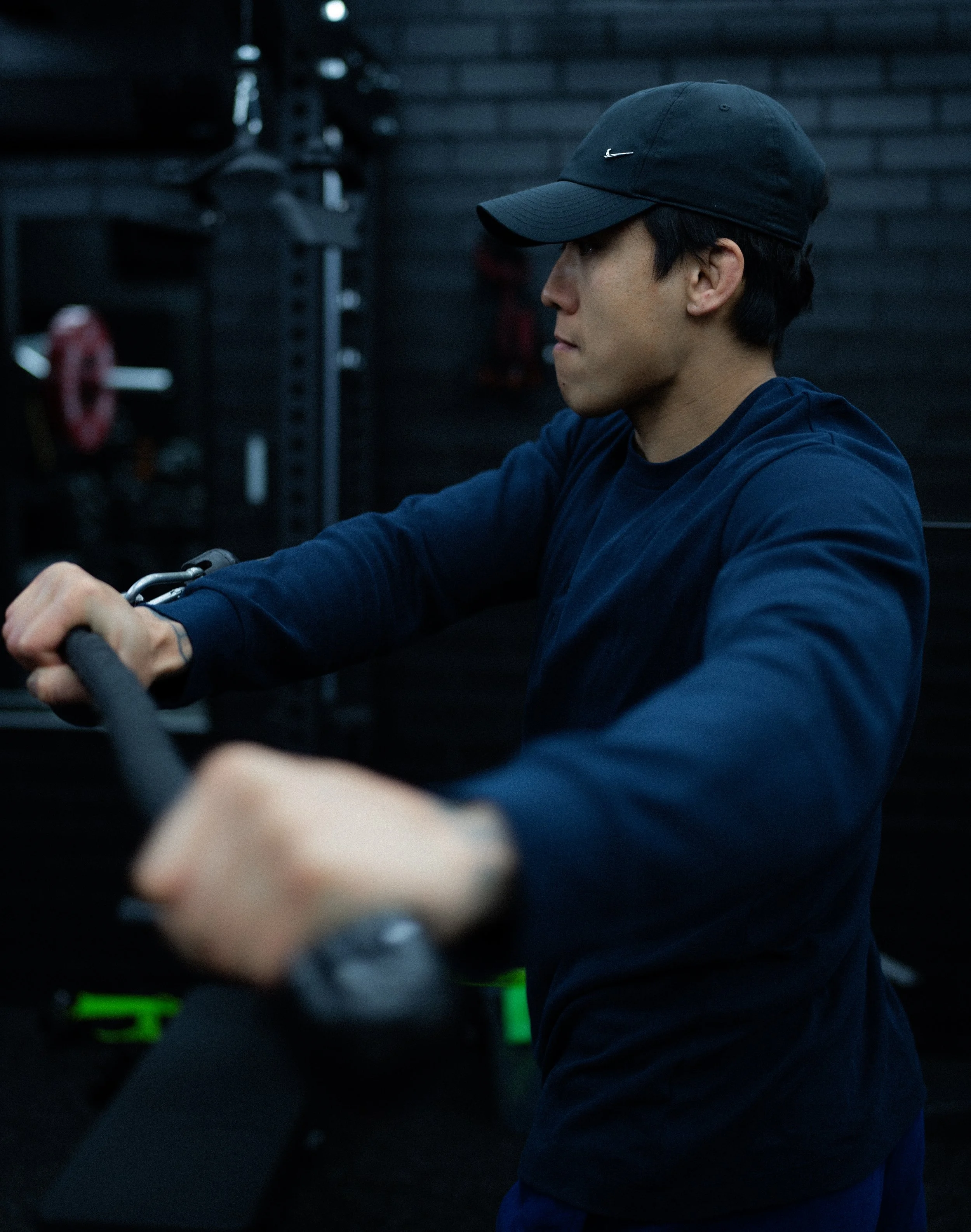 A man wearing a black Nike cap and a dark blue long-sleeve shirt is working out with a resistance band in a dimly lit gym. He is holding the band with both hands, and the background includes gym equipment and a dark wall.