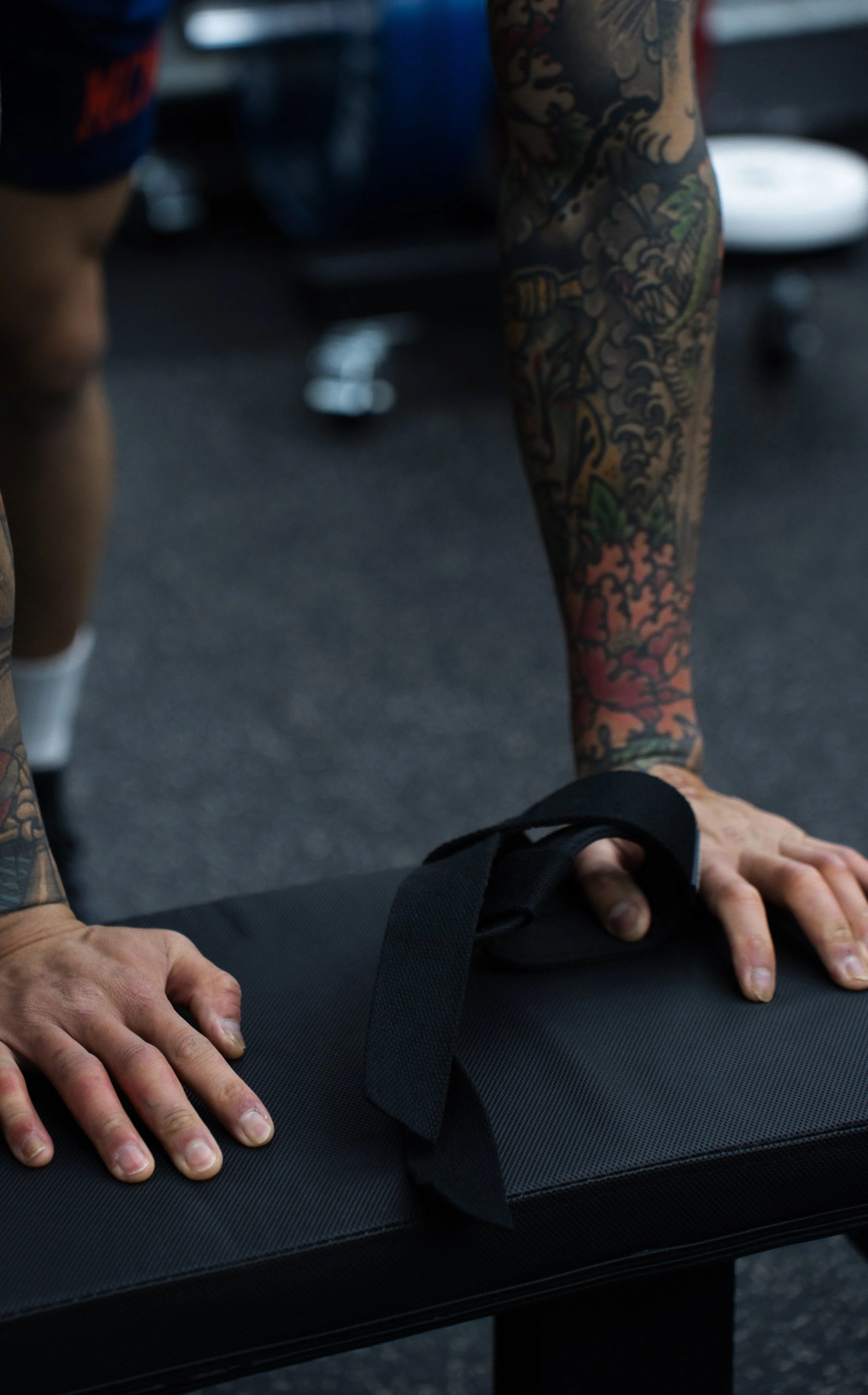 Person with tattoos on arms and hands, placing hands on a black workout mat, with exercise equipment in the background.