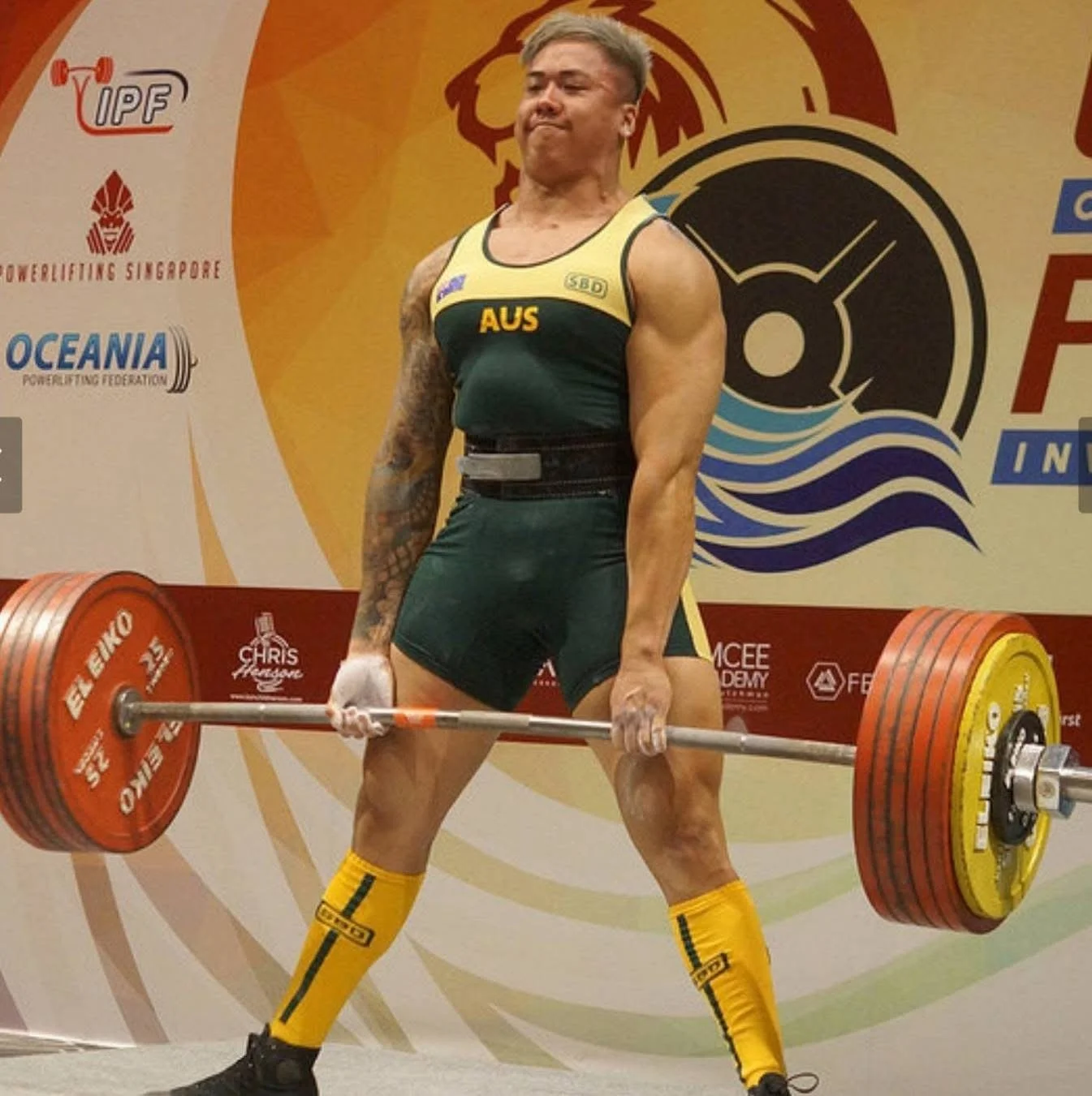 A male weightlifter wearing a green and yellow Australia uniform, lifting a heavy barbell with red and yellow weights, wearing yellow socks and black shoes, with a backdrop featuring logos and text related to powerlifting events.