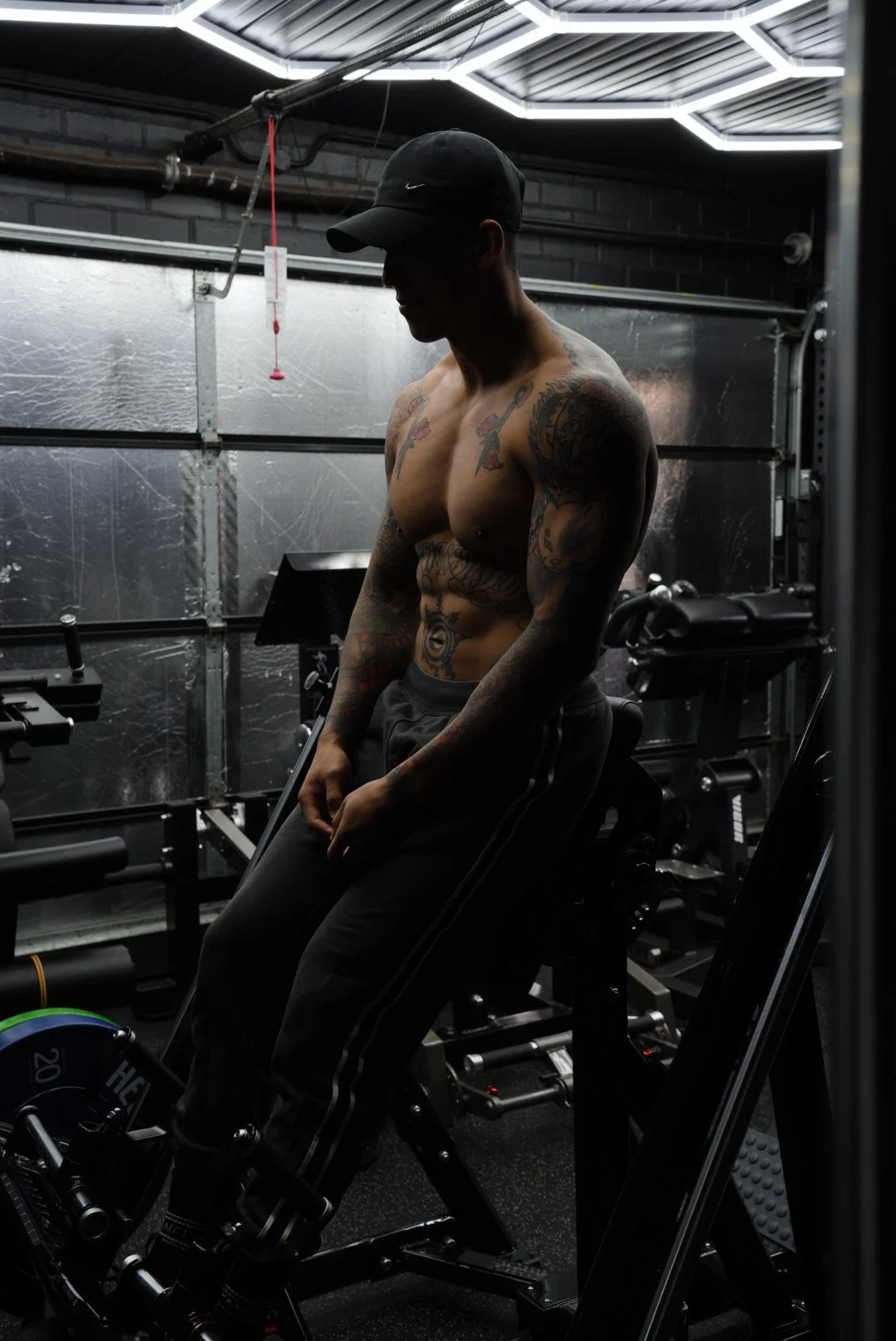 A shirtless man with tattoos, wearing a cap, sitting in a gym surrounded by workout equipment and illuminated by modern ceiling lights.