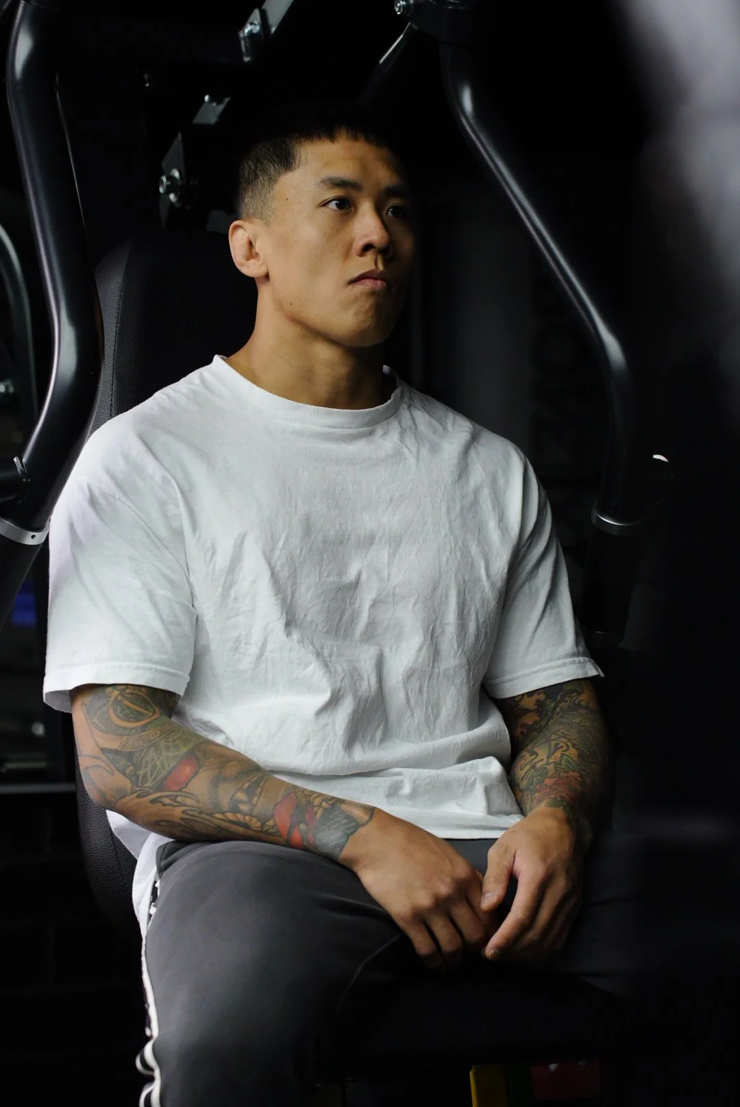 A young man with short black hair and tattoos on his arms sitting in a chair, wearing a white t-shirt.