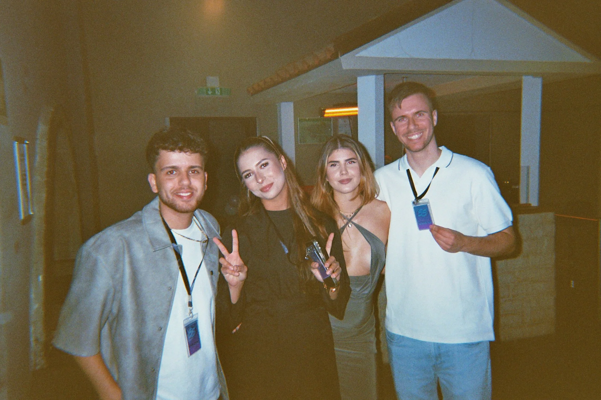 Vier junge Menschen, drei Männer und eine Frau, stehen zusammen in einer dunklen, indoor Umgebung und posieren für die Kamera, einige halten Gegenstände, zwei der Frauen machen Friedenszeichen.