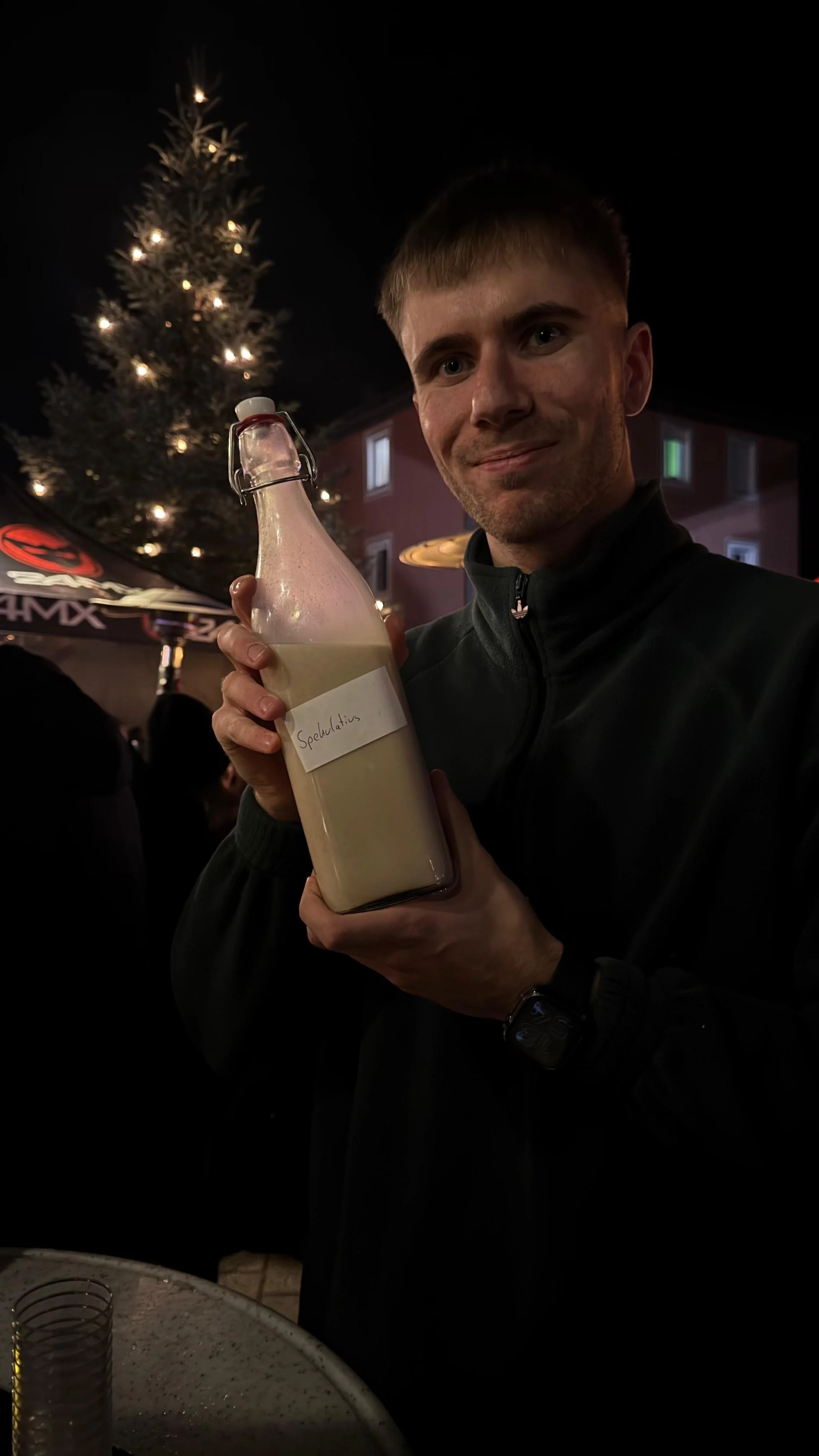 Ein Mann hält eine Flasche mit einer weißen Flüssigkeit, die beschriftet ist mit "Spekulatius", bei einer Feier im Freien, im Hintergrund ist ein dekorierter Weihnachtsbaum mit Lichtern sichtbar.