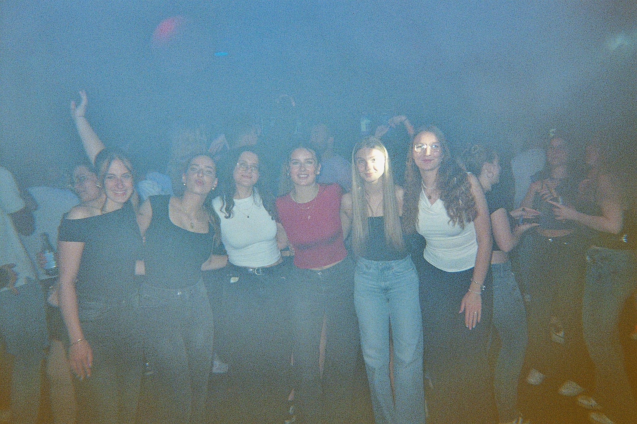 Sechs junge Frauen stehen zusammen in einer dunklen, verrauchten Party-Umgebung, lächeln und posieren für die Kamera.