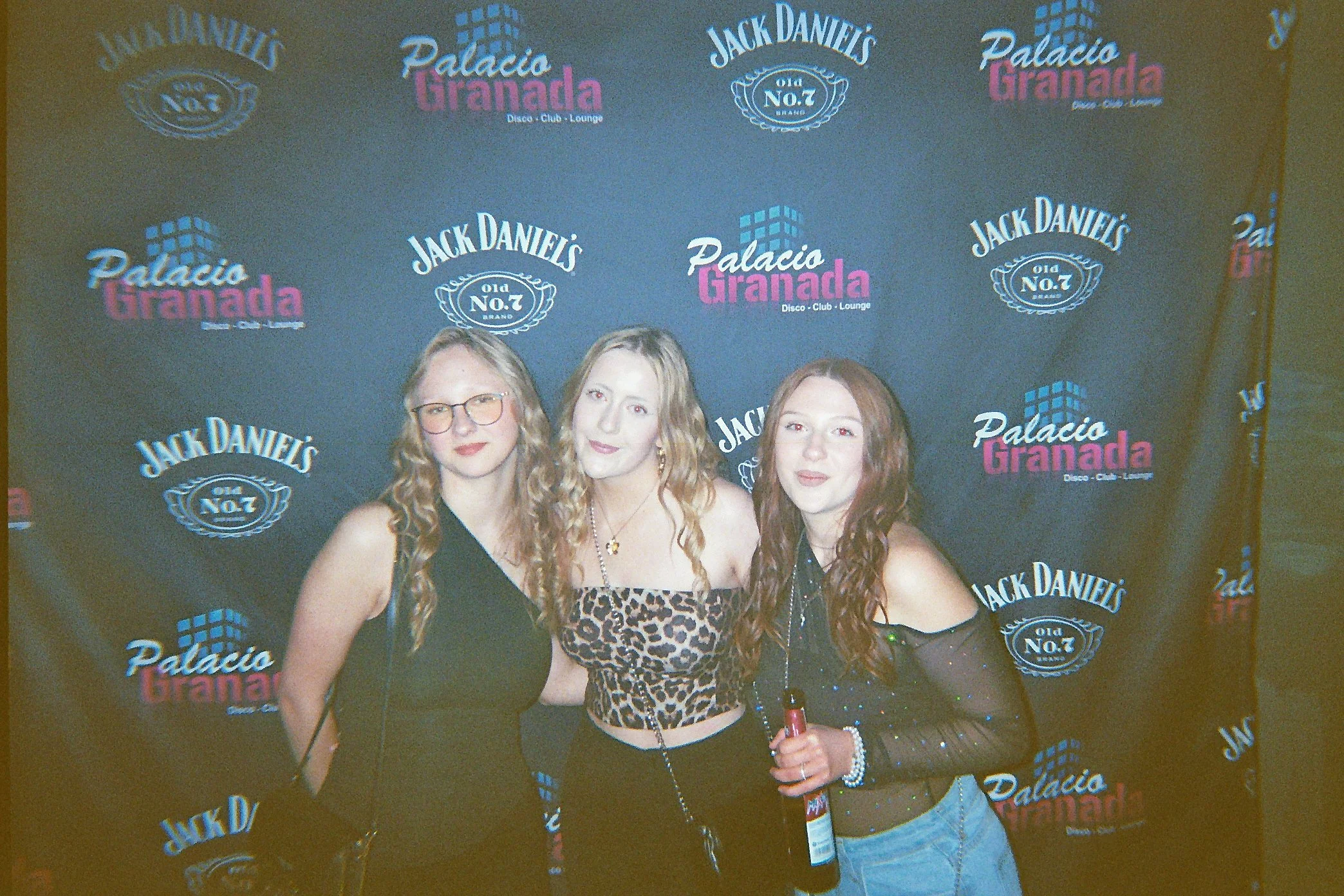 Drei Frauen posieren vor einem schwarzen Hintergrund mit dem Logo "Palacio Granada" und "Jack Daniel's". Sie tragen schwarze Outfits; die Frau in der Mitte trägt ein leopardmusterndes Top.