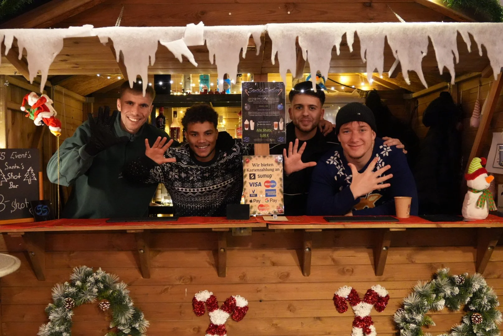 Vier junge Männer stehen hinter einem Weihnachtsstand, der mit Weihnachtsdekor dekoriert ist, und winken in die Kamera.