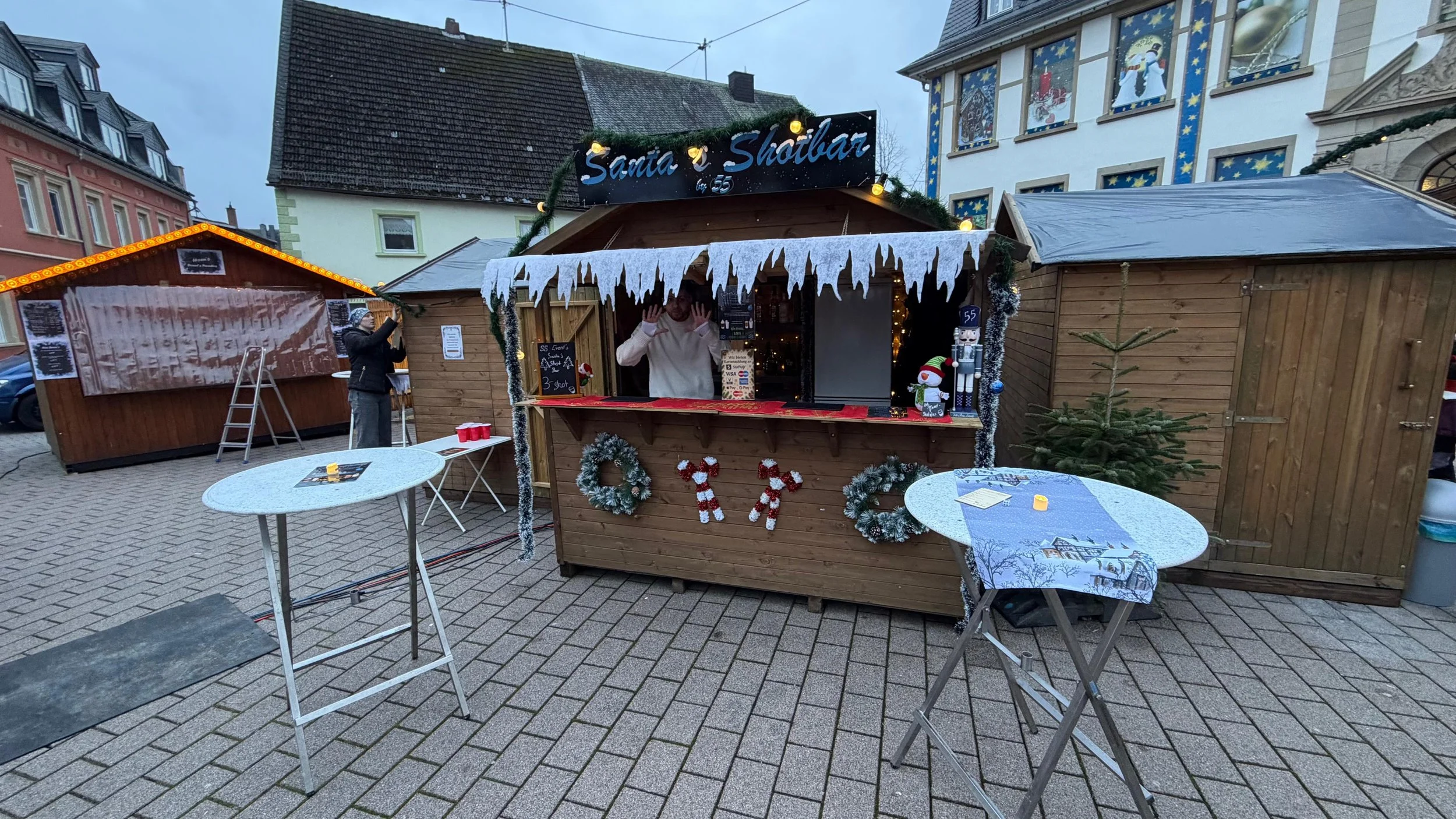 Weihnachtsmarktstand mit Dekorationen, Tannenbaum, und Weihnachtsmotive, im Hintergrund Gebäude mit Weihnachtsbildern, draußen auf gepflastertem Platz.