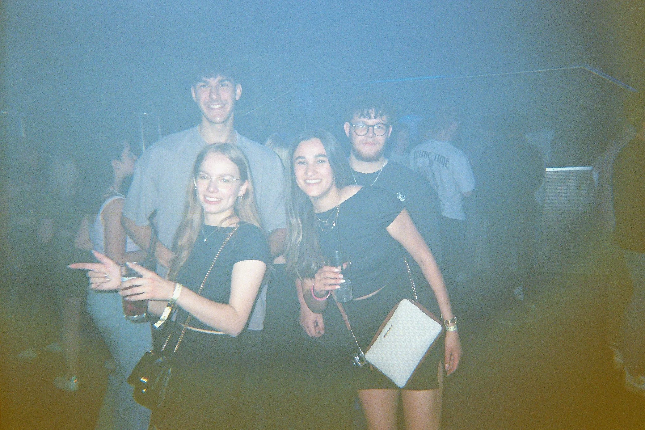 Vier junge Menschen feiern in einer Bar oder einem Club, zwei Frauen und zwei Männer, lachen und halten Getränke, die Atmosphäre ist lebhaft.