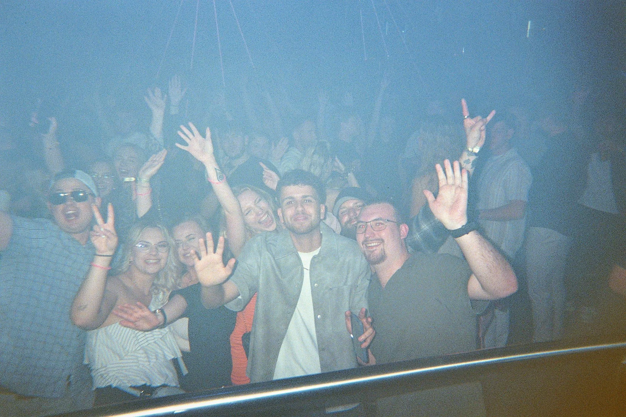 Gruppen von jungen Menschen feiern in einer dunklen, verrauchten Partyumgebung, einige sind winkend und lächelnd in die Kamera.