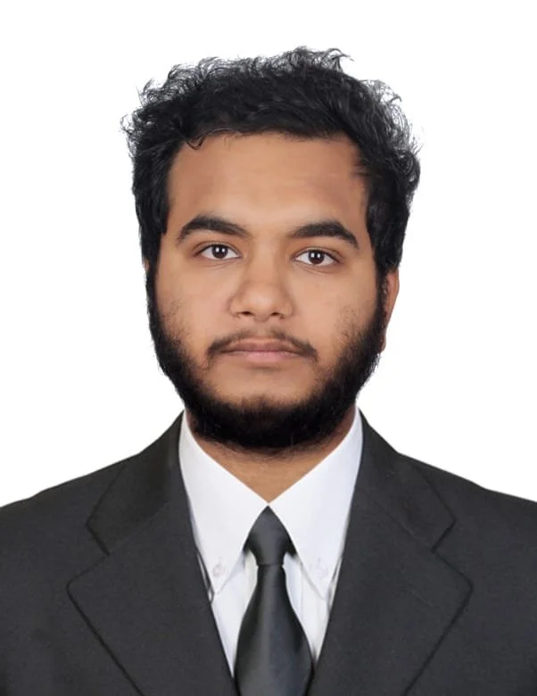 Portrait of a young man with curly black hair and a beard, wearing a black suit, white shirt, and black tie, against a plain white background.