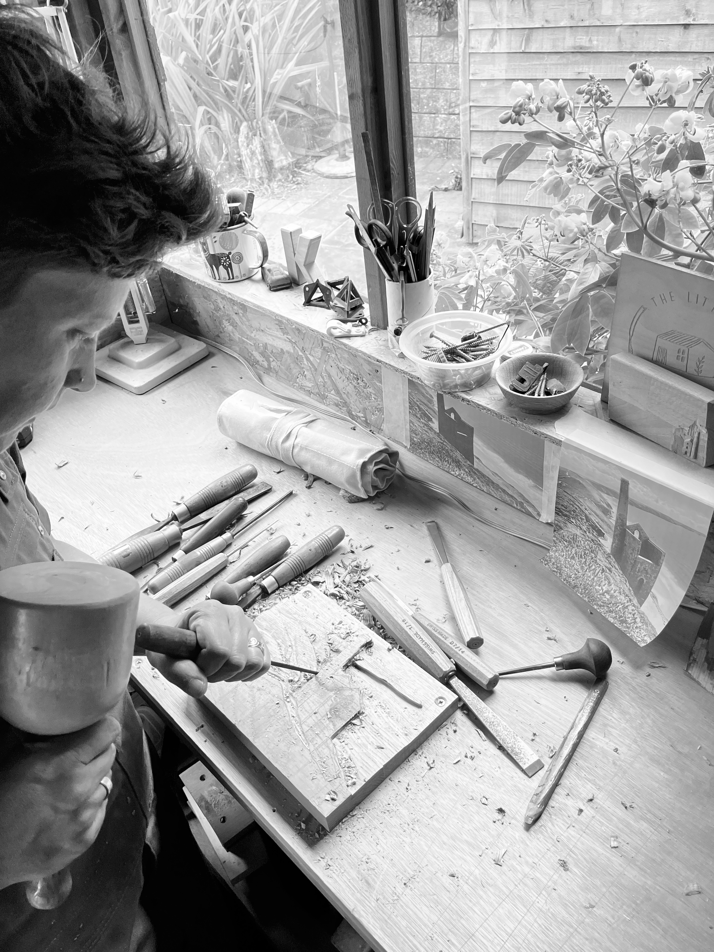 A person working on a woodworking project in a workshop, using a mallet and chisel on a piece of wood, with various woodworking tools and supplies on the workbench and window ledge.