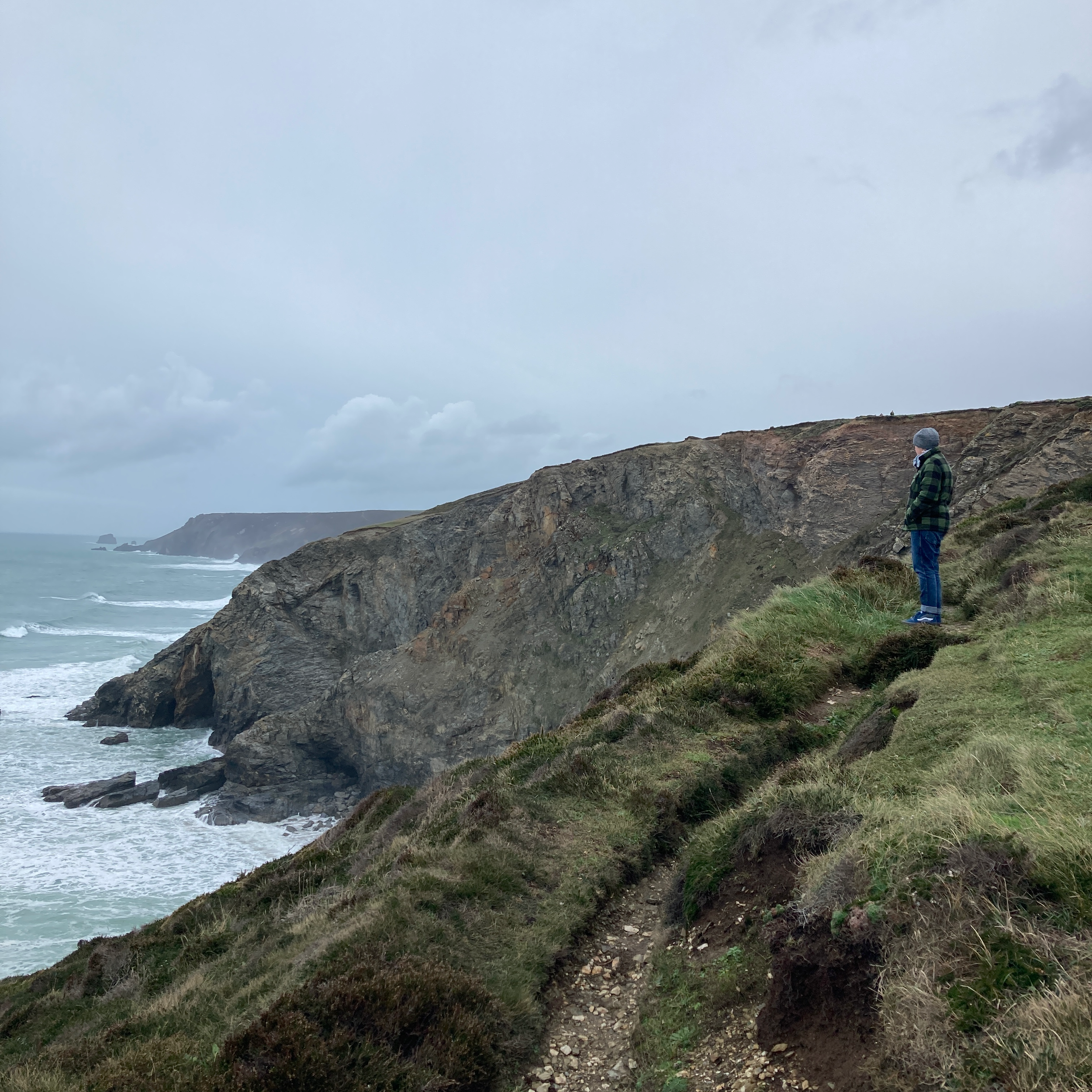A person standing on a grassy cliffside looking out towards the ocean, with rocky cliffs in the distance and cloudy skies above.