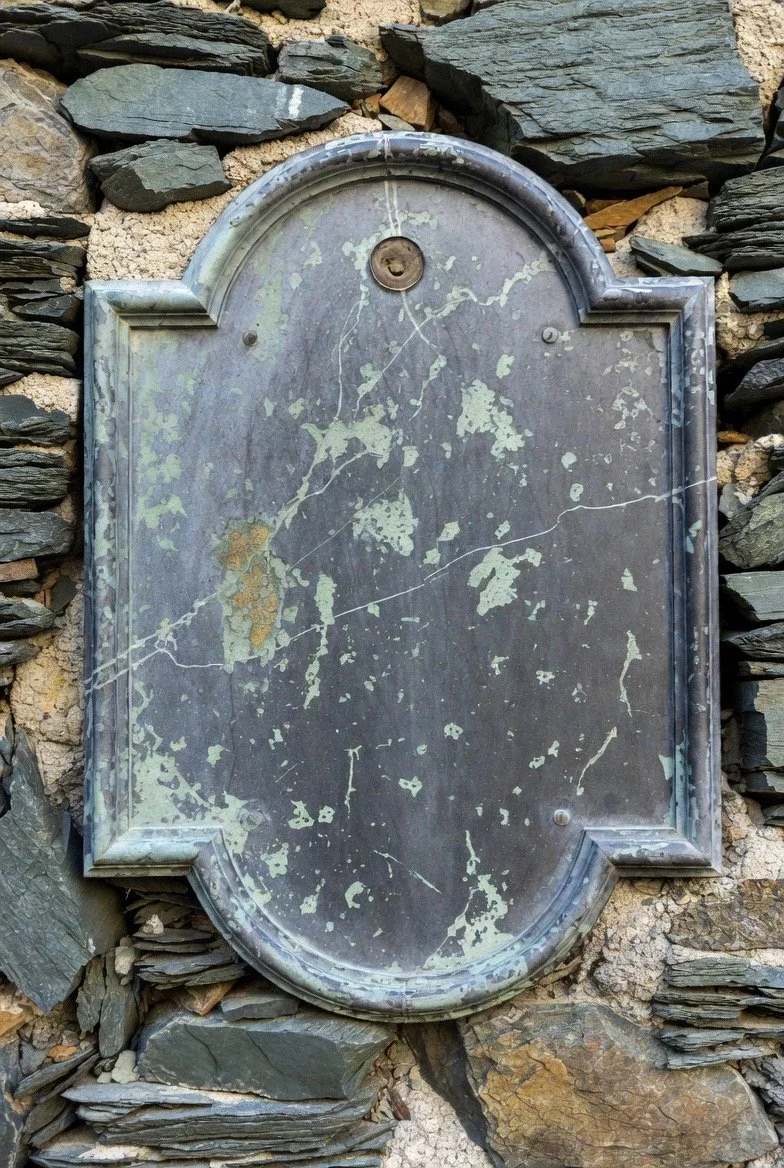 Weathered metal plaque with peeling paint, mounted on a stone wall, with a small lock or keyhole at the top center.
