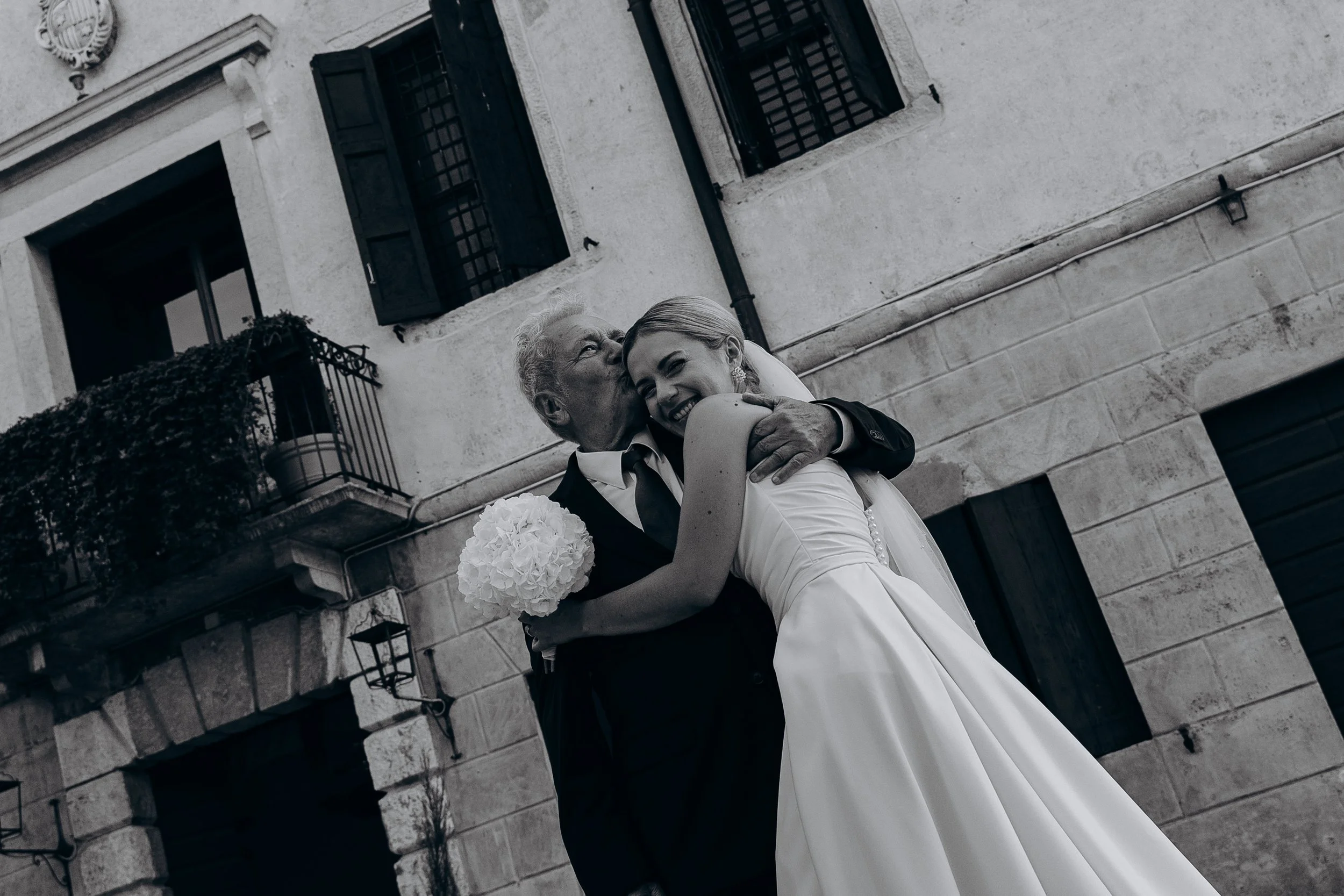 Un matrimonio abbraccia una donna anziana, la scena è all'aperto davanti a un edificio con finestre con sbarre e un balcone con piante in vaso.
