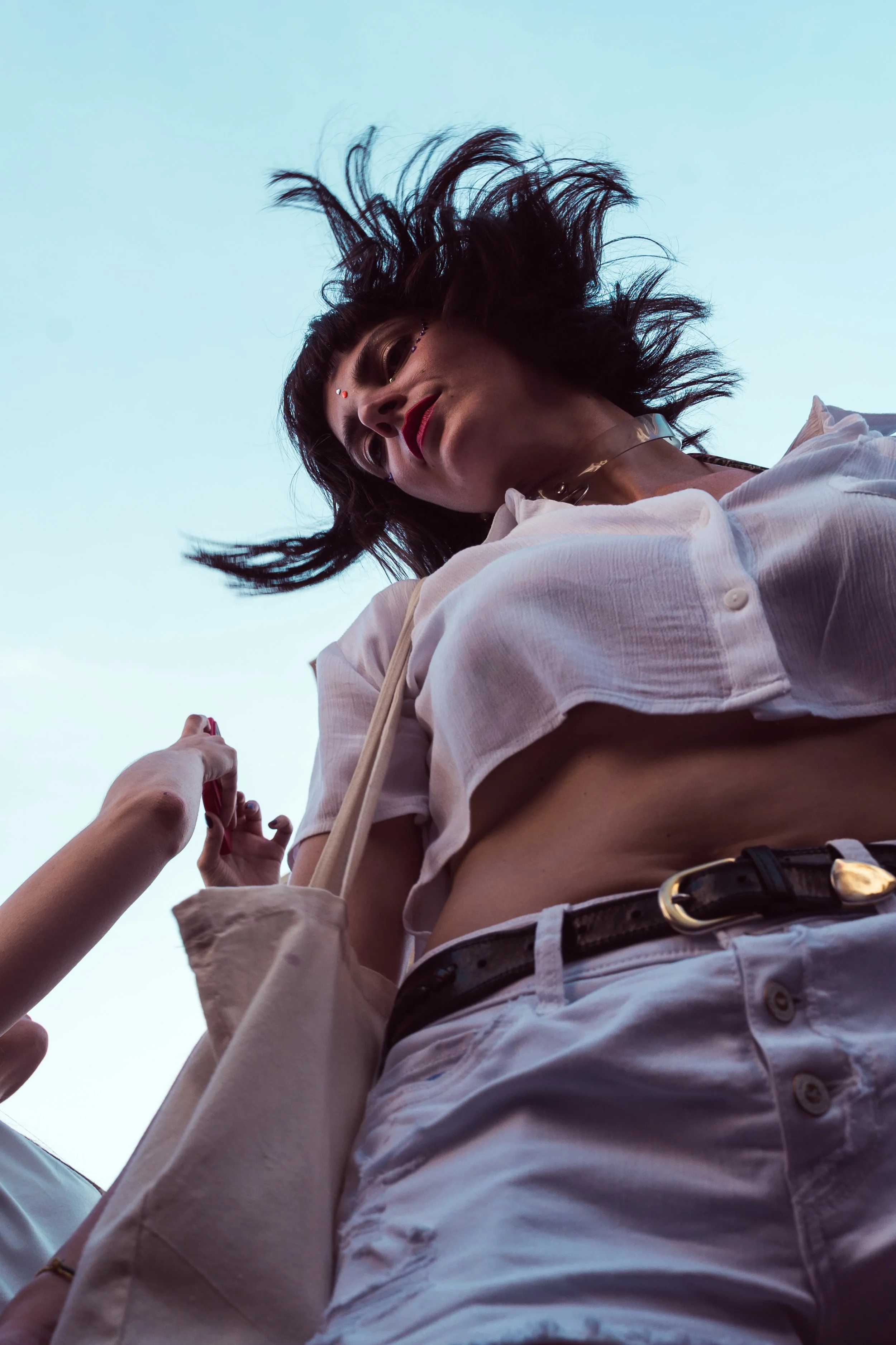 Una donna con cappelliere e capelli mossi indossa una camicia bianca e pantaloni bianchi, con una cintura nera, mentre qualcuno le tocca la tasca sul lato sinistro.