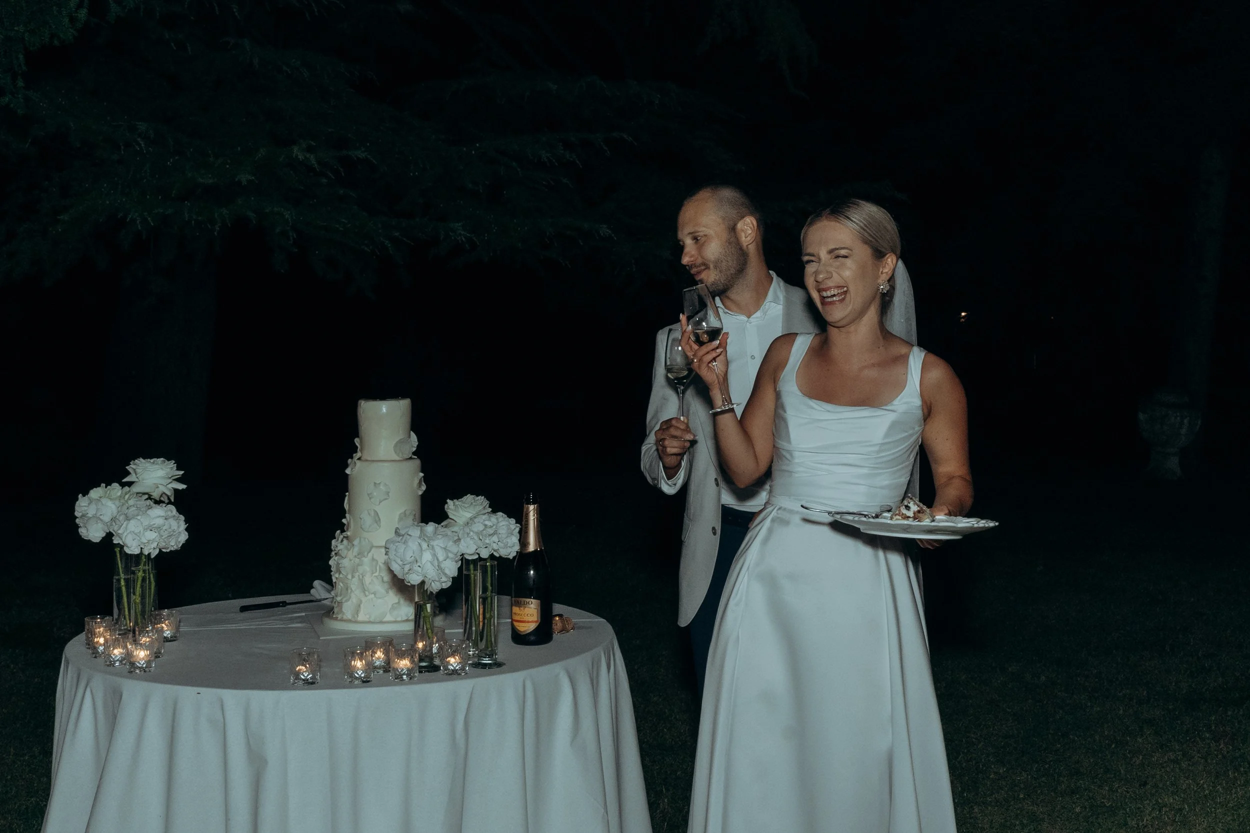 Coppia di sposi con abiti bianchi che brindano durante il ricevimento di nozze all'aperto di sera, con torta nuziale e decorazioni floreali sulla tavola.