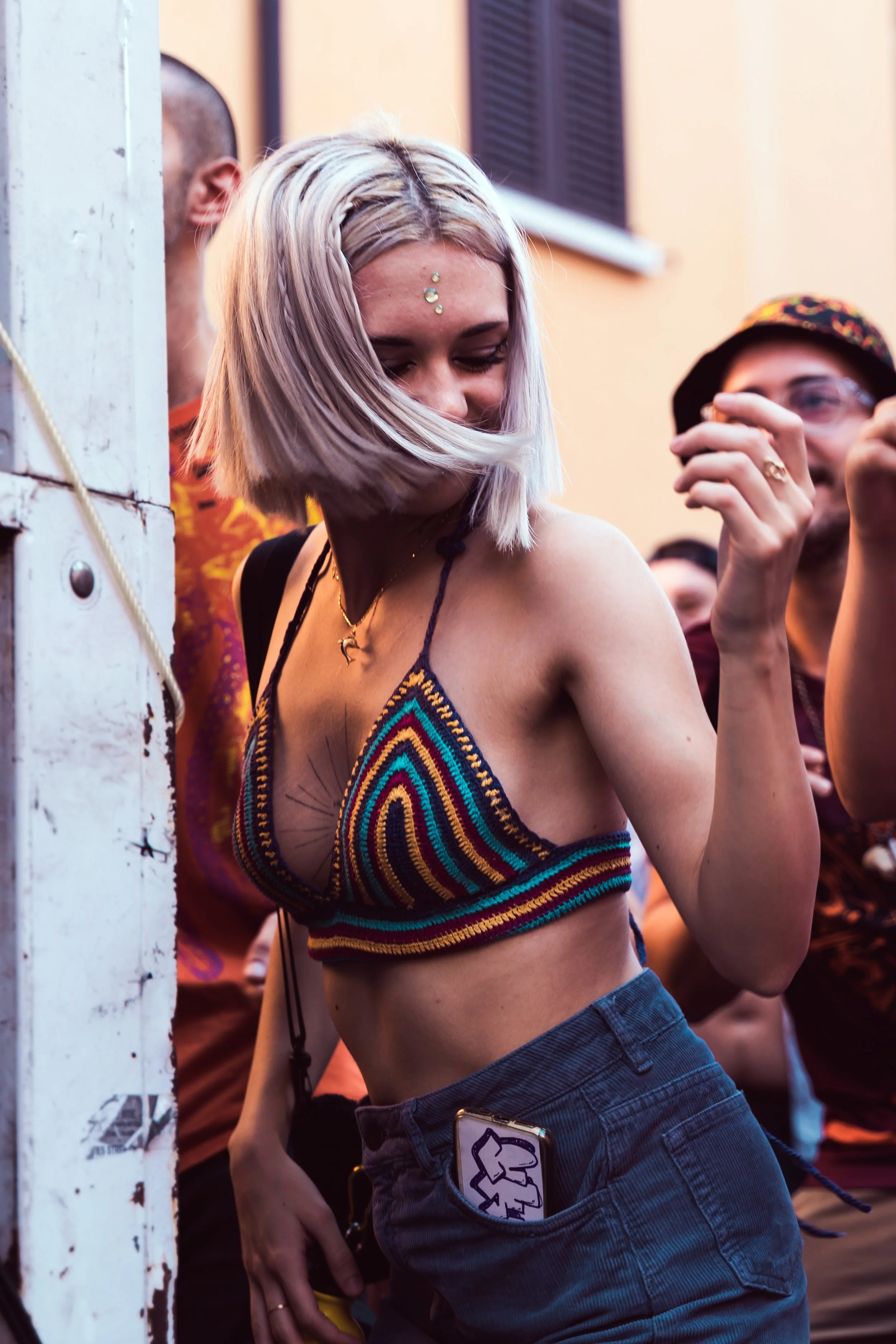 Una donna con capelli biondo platino, vestita con un top regular a rete colorata, si trova tra un gruppo di persone che sembrano divertirsi, in un contesto di festa o evento sociale.