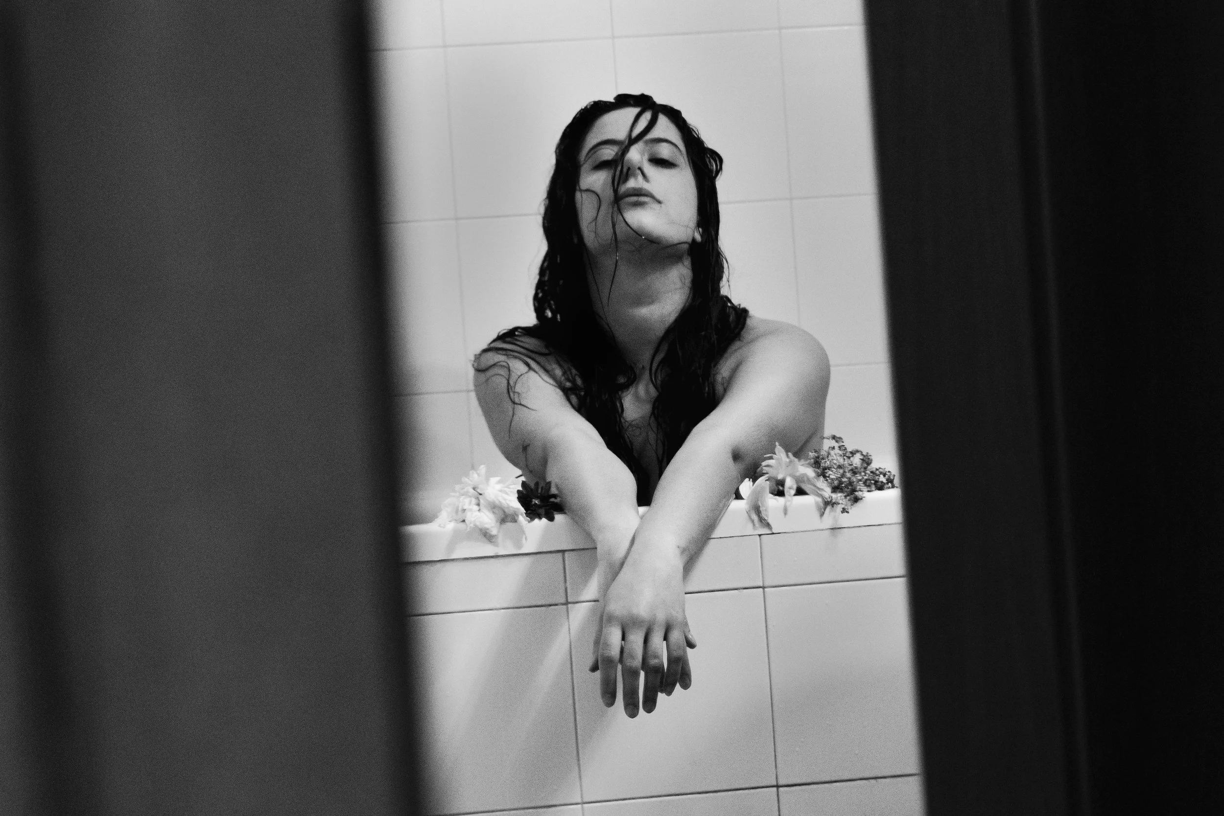 Una donna con i capelli bagnati, affiora da una vasca da bagno circondata da fiori, vista tramite uno specchio o una porta aperta, in un ambiente di colore bianco e nero.