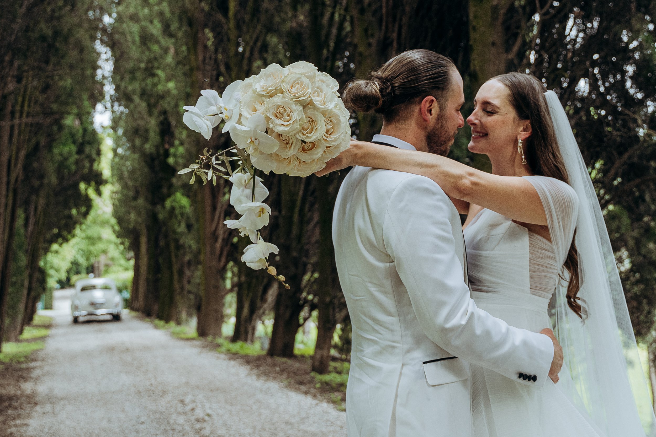 Una coppia di sposi abbracciati davanti a un albero, con una strada sterrata e una macchina in lontananza, nel contesto di un matrimonio all'aperto.