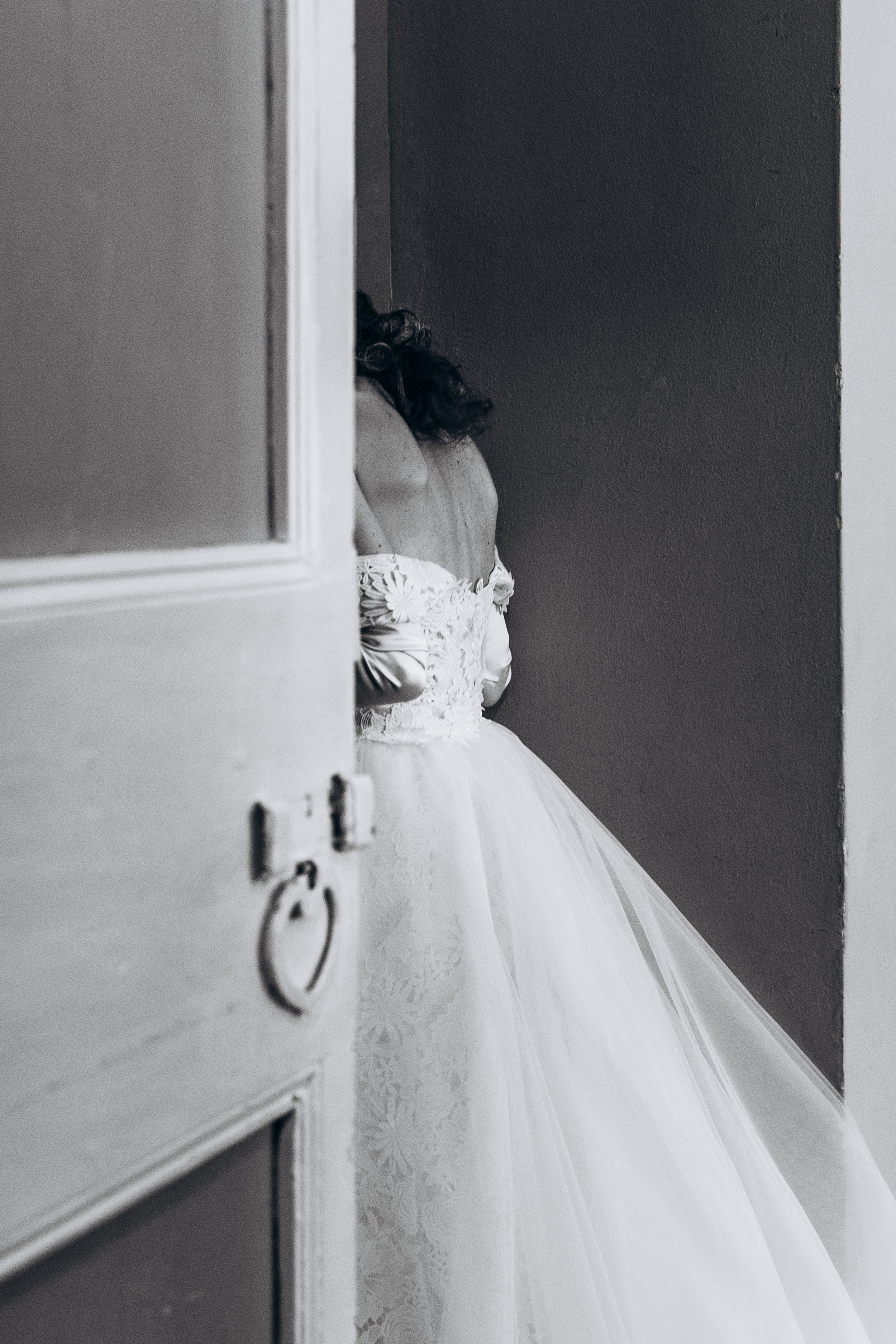 Donna in abito da sposa dietro una porta semiaperta, visibile solo la schiena e parte dei capelli, in una stanza con pareti scure.