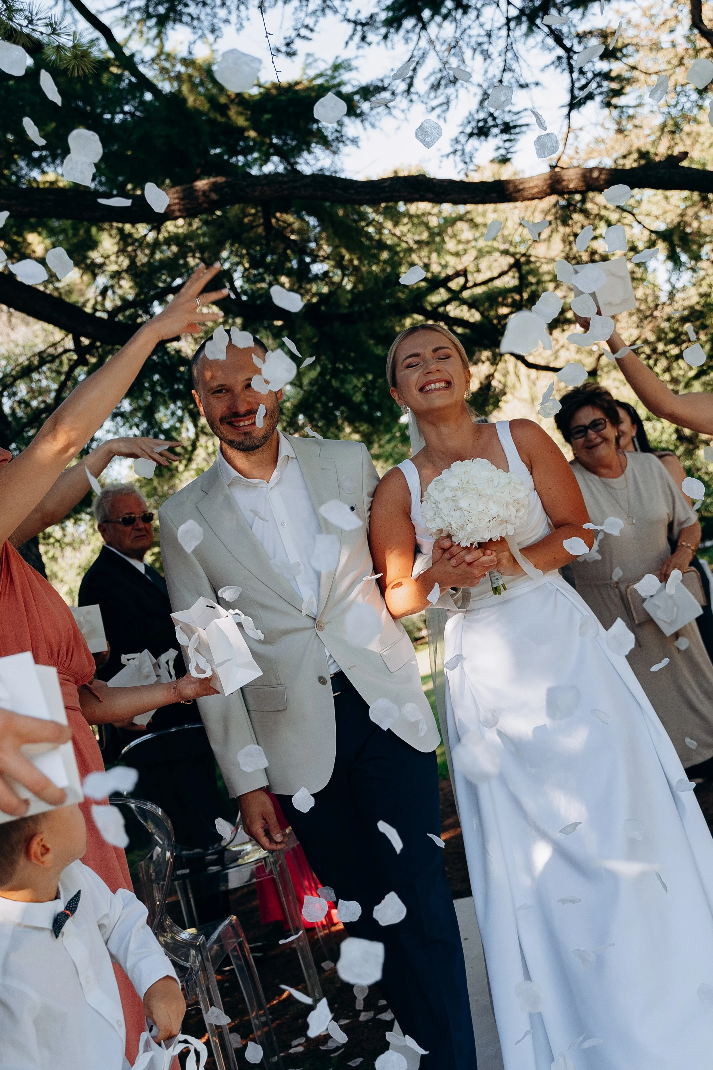 Sposi di nozze che camminano sotto coriandoli bianchi, circondati da amici e familiari in un matrimonio all'aperto sotto un albero.