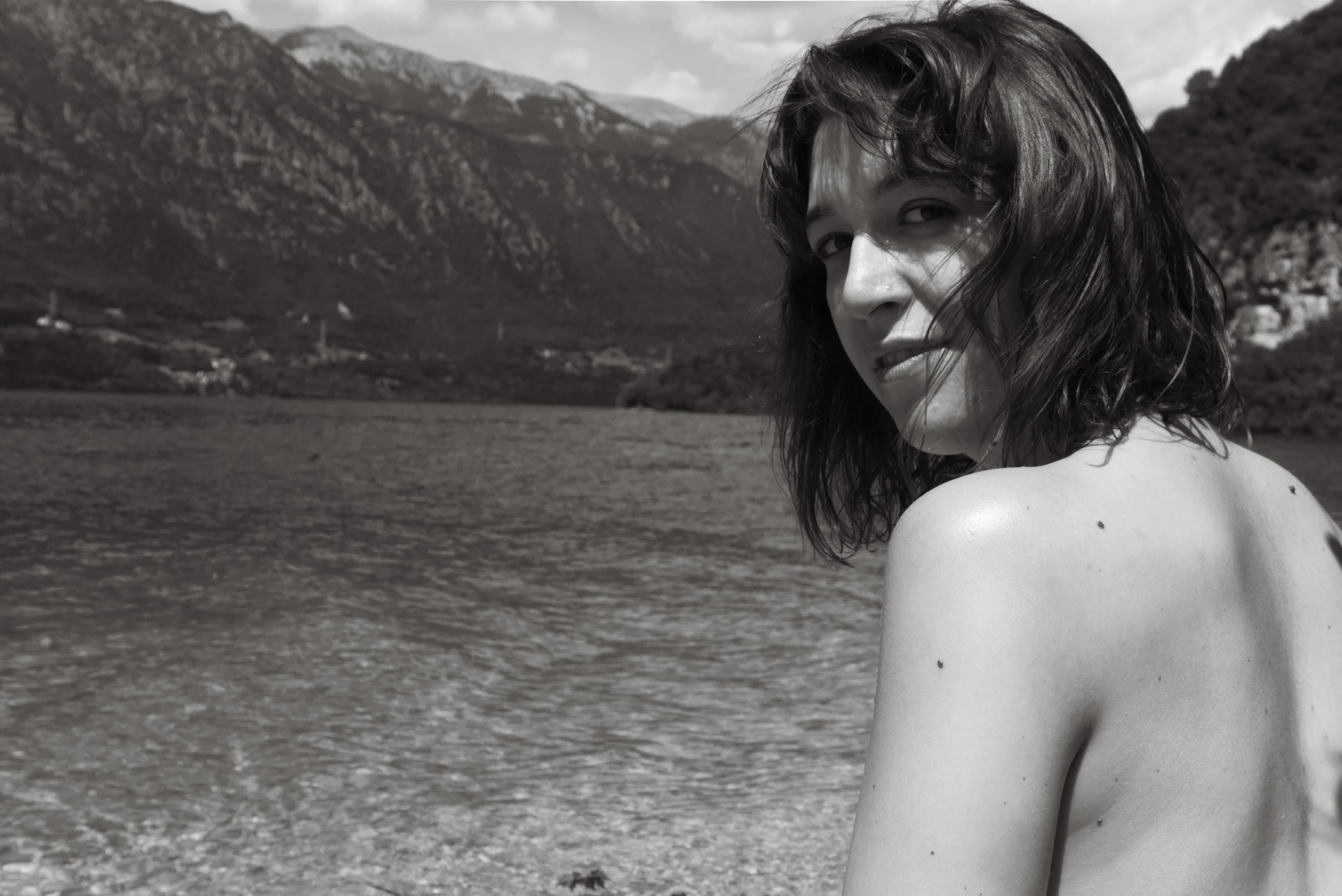 Ragazza con capelli mossi, vista di spalle, siede vicino a un lago con montagne sullo sfondo, in una fotografia in bianco e nero.