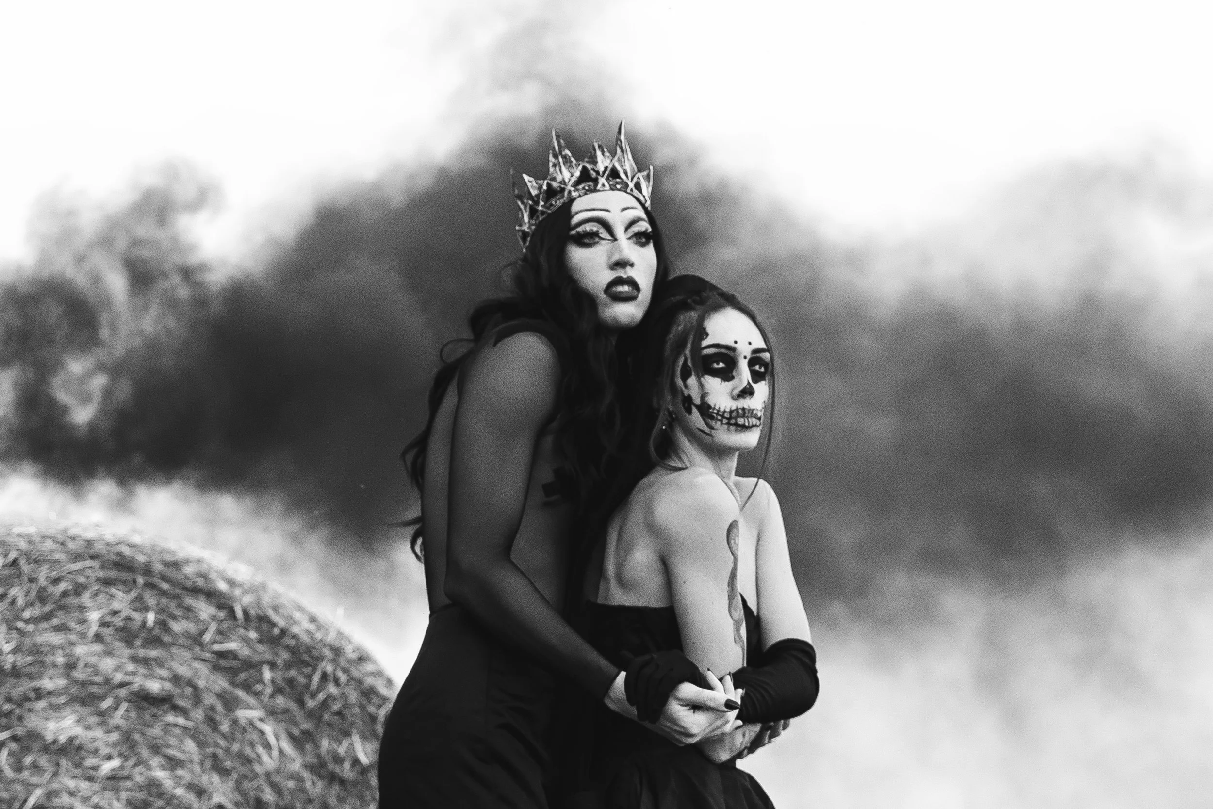 Due donne con trucco e costumi artistici, una indossa una corona e l'altra un trucco scheletrico, in un paesaggio con nuvole scure sullo sfondo