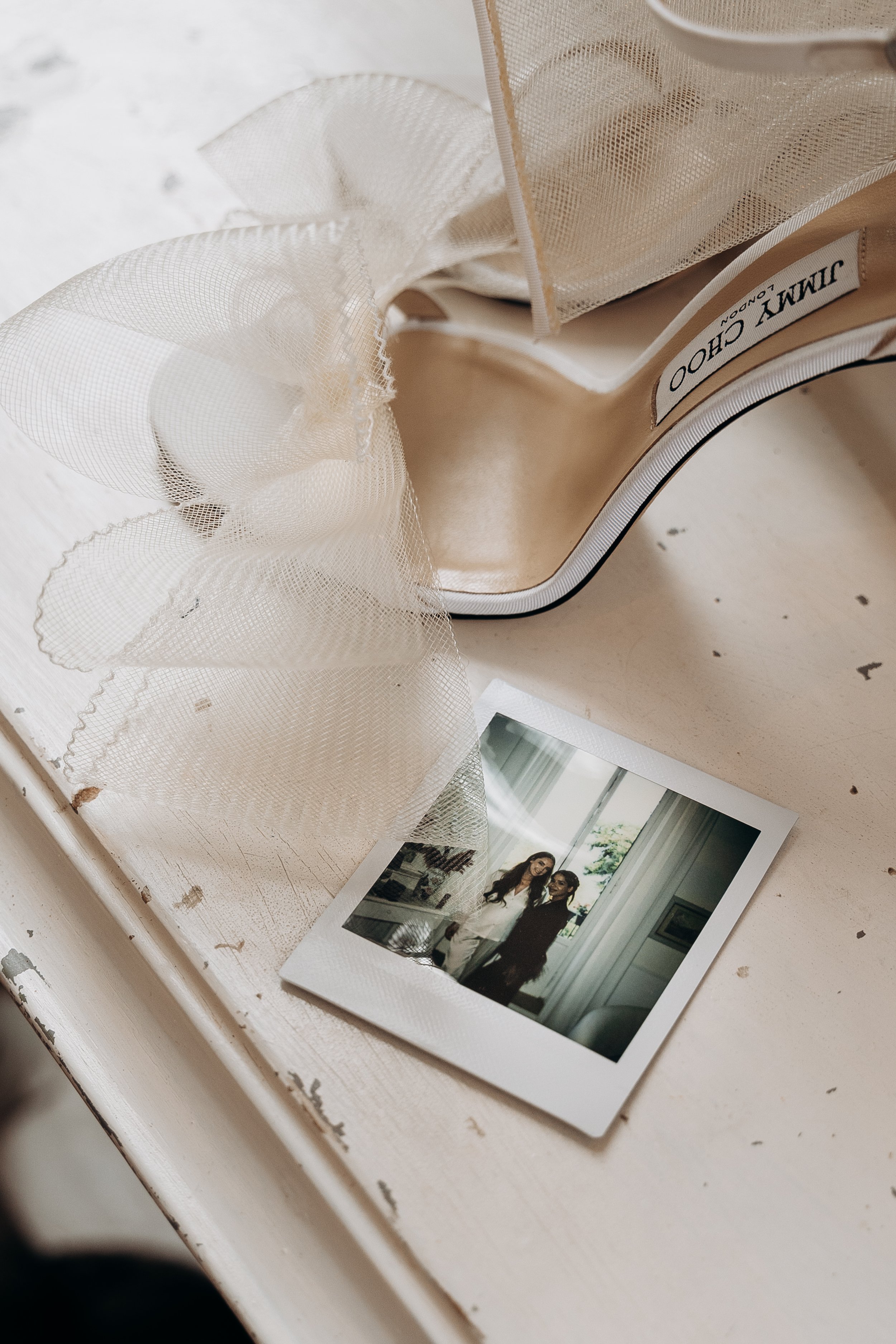 Una scarpa da sera beige con dettagli di tulle, una fotografia polaroid di due donne sorridenti e un tavolo di legno bianco leggermente usurato.