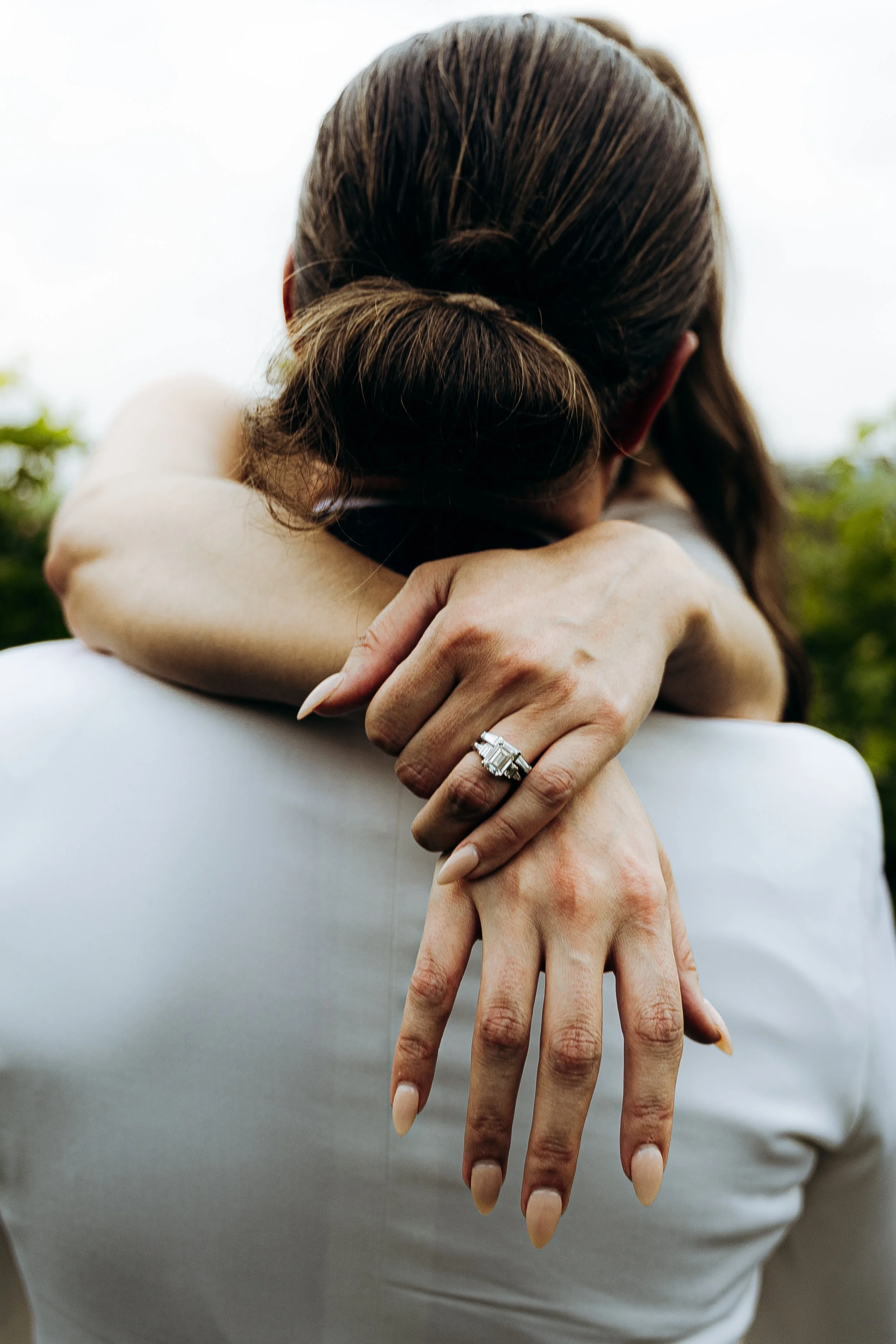 Due persone che si abbracciano, una delle quali mostra un anello di diamanti su una mano, con una scena naturale sfocata sullo sfondo.