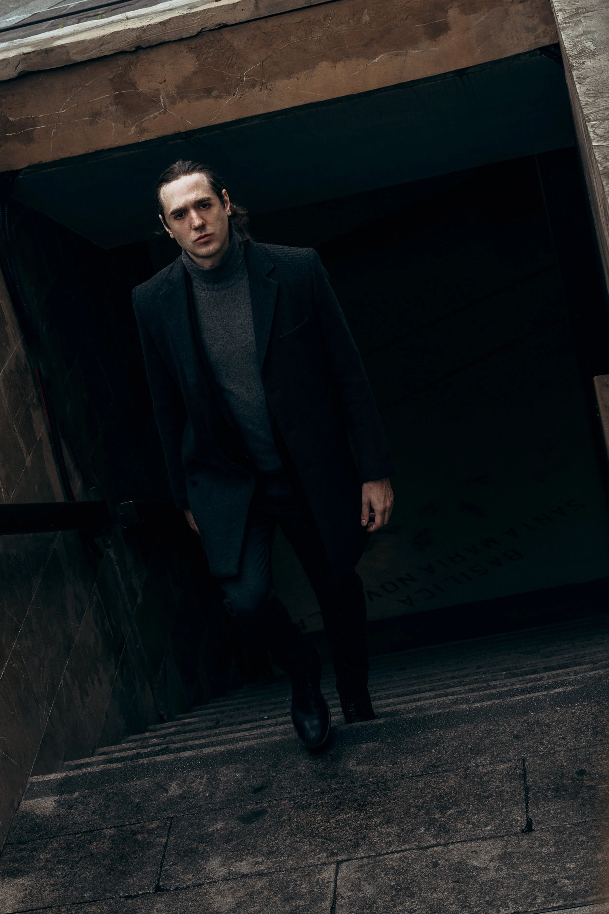 Uomo in abbigliamento scuro che sale le scale di una struttura con pareti di pietra, in un'illuminazione soffusa.