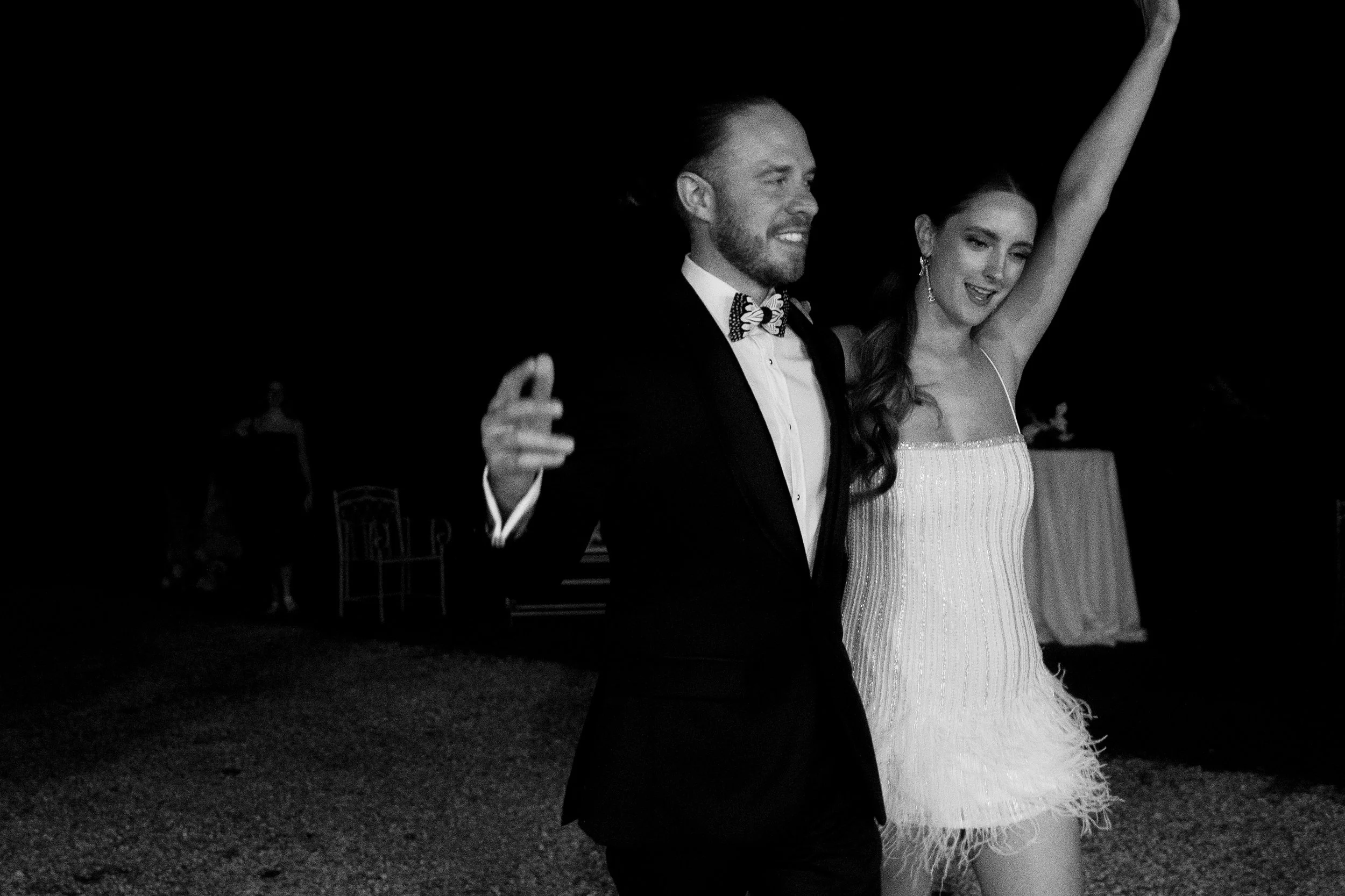 Una coppia di sposi balla e sorride durante il loro matrimonio, l'uomo indossa uno smoking con papillon, la donna indossa un vestito da sposa corto con dettagli scintillanti, sullo sfondo atmosfera festiva.